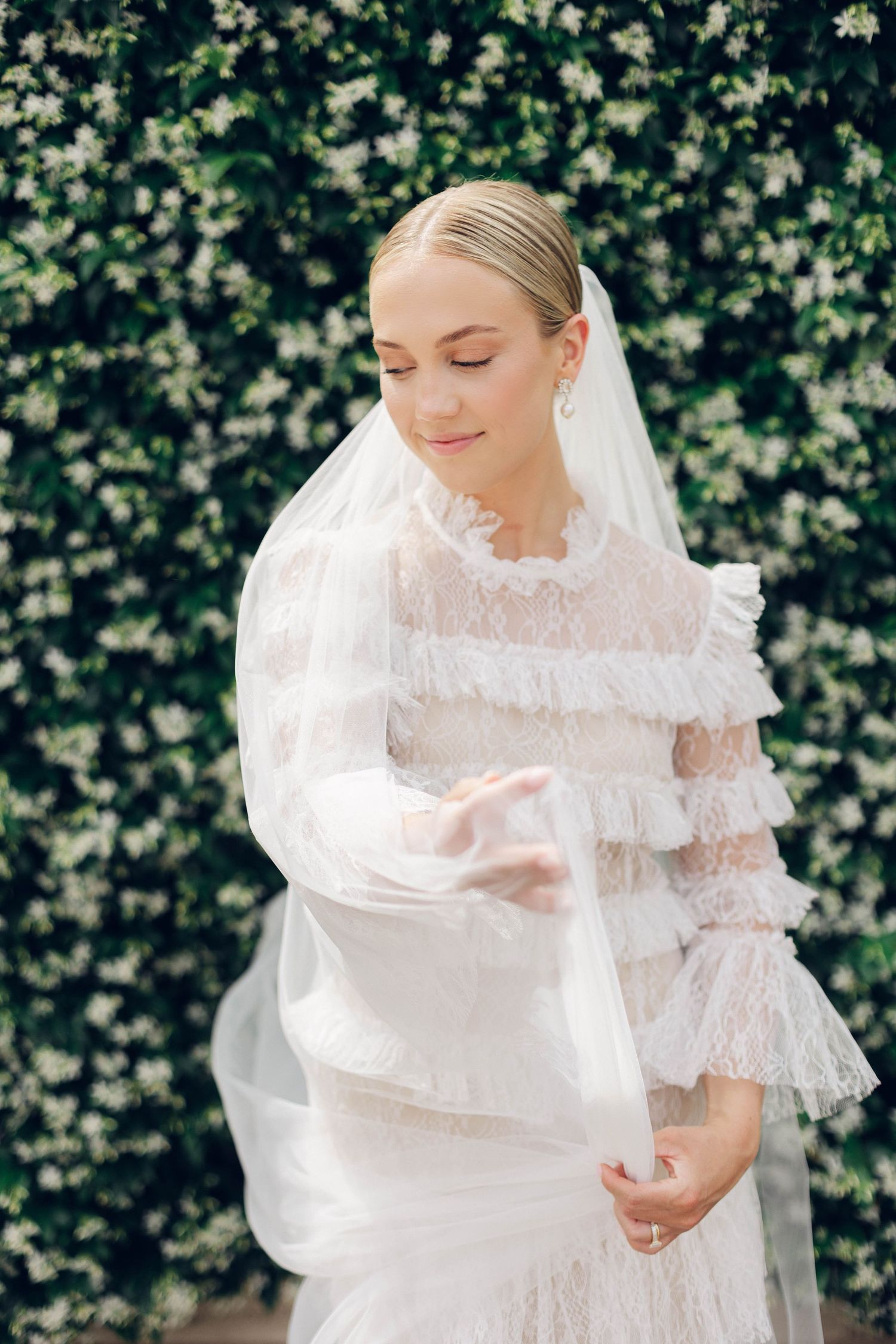 Bride in layered lace dress with veil against flowering wall background.