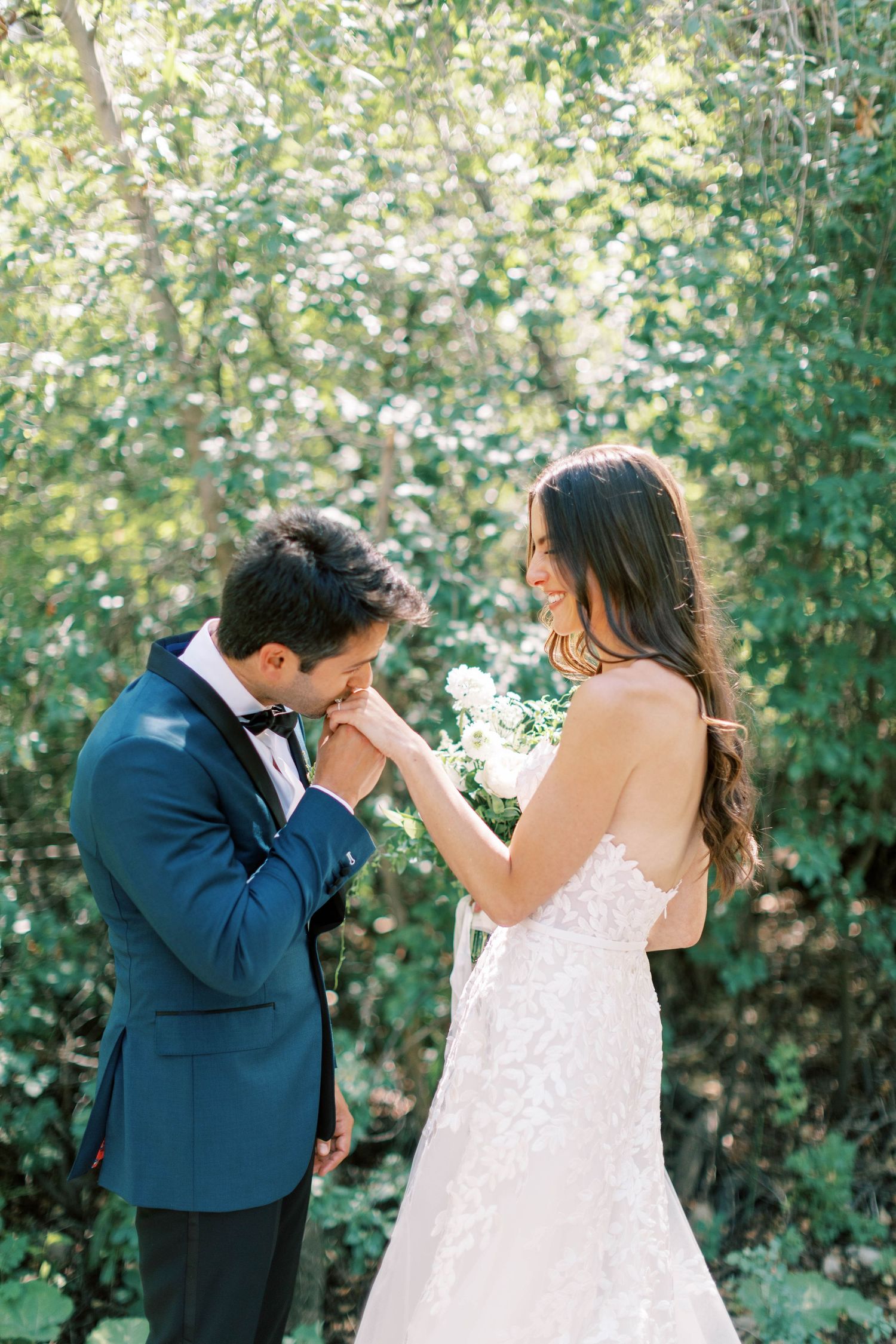Groom in blue suit kisses bride's hand during outdoor first look moment.