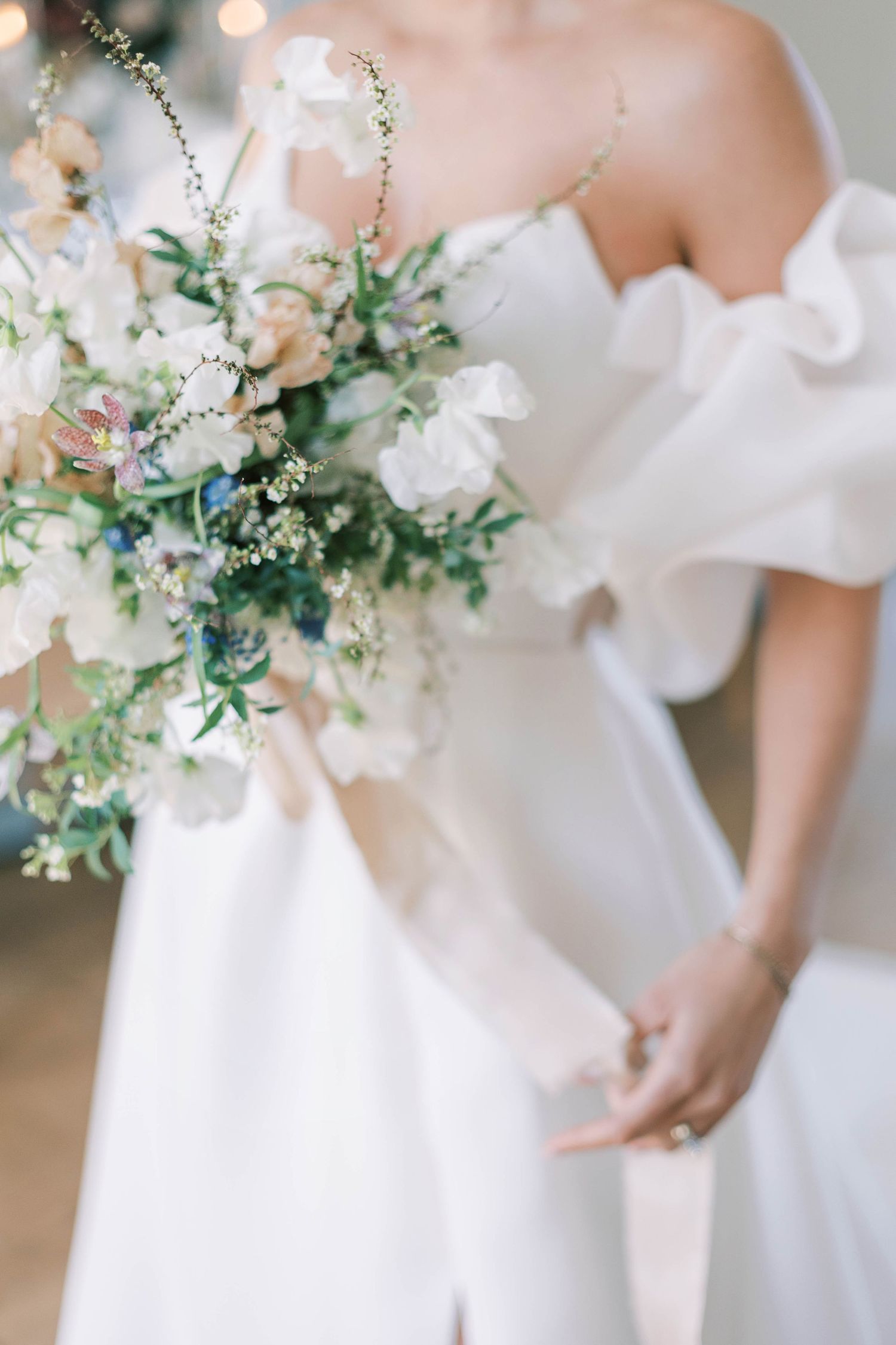 Delicate white wedding bouquet with greenery shown against off-shoulder bridal gown.