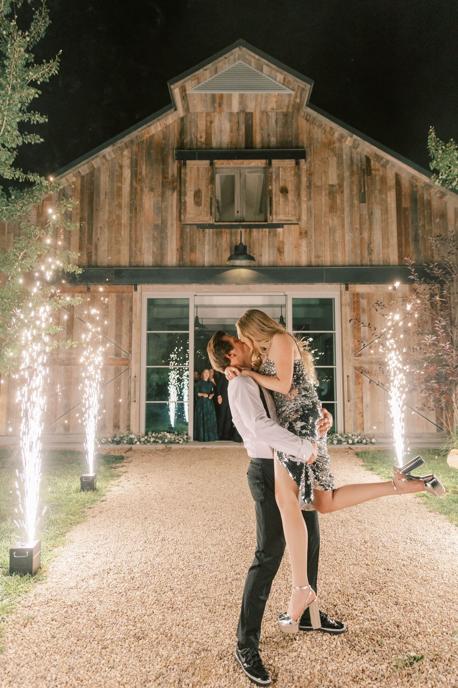 Couple sharing intimate moment in front of barn wedding venue lit by sparkling fountains.