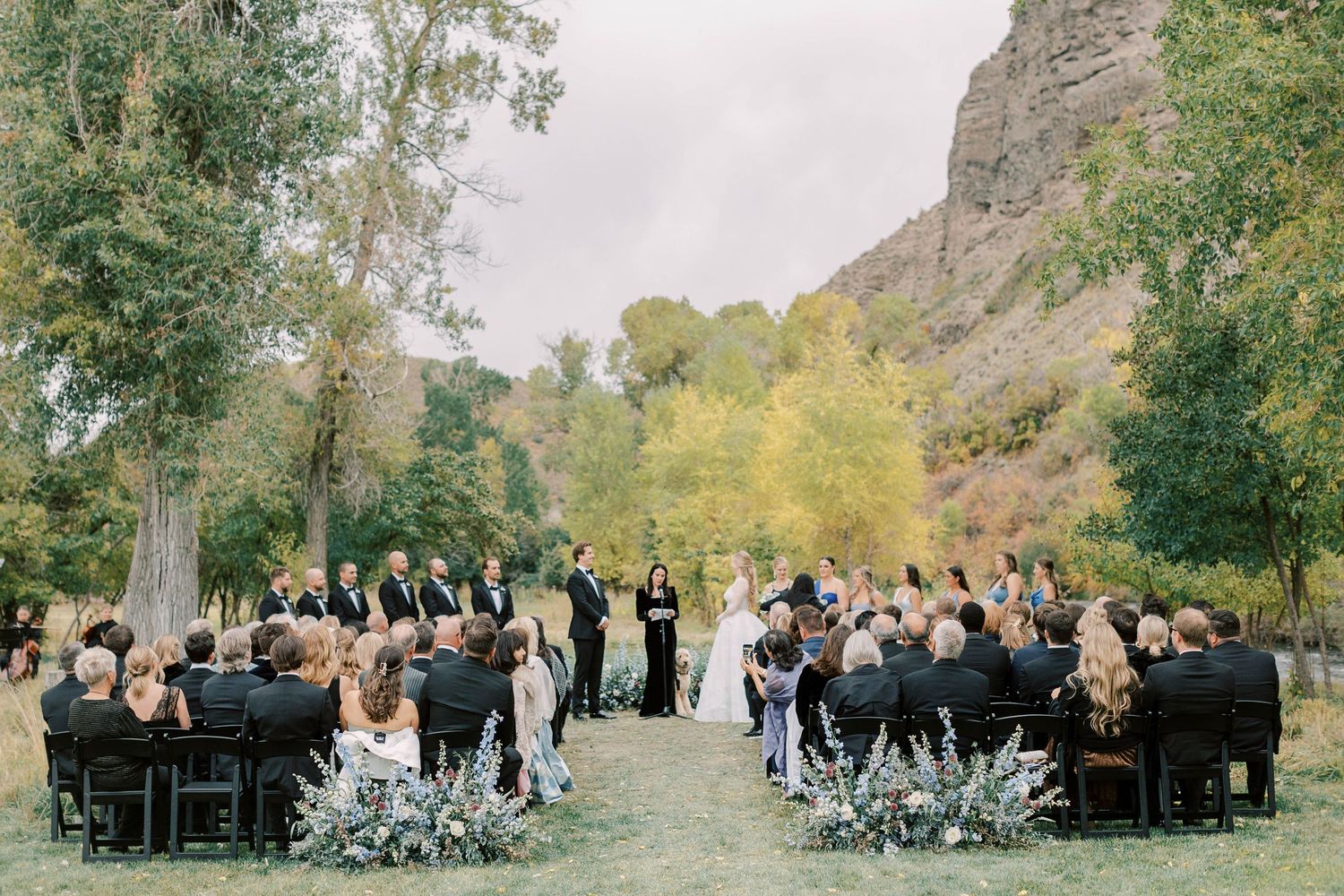 Overview of outdoor wedding ceremony with guests seated beneath towering cliffs and autumn trees.