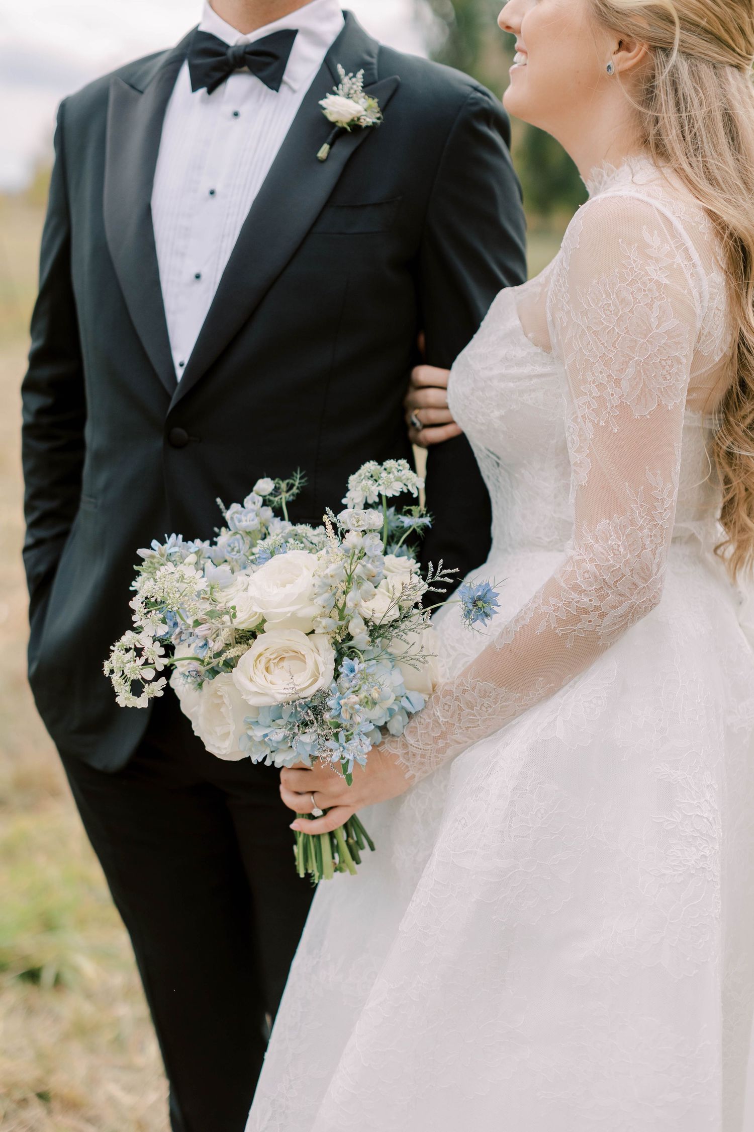 Close-up of bride holding delicate white and blue bouquet next to groom in classic tuxedo.
