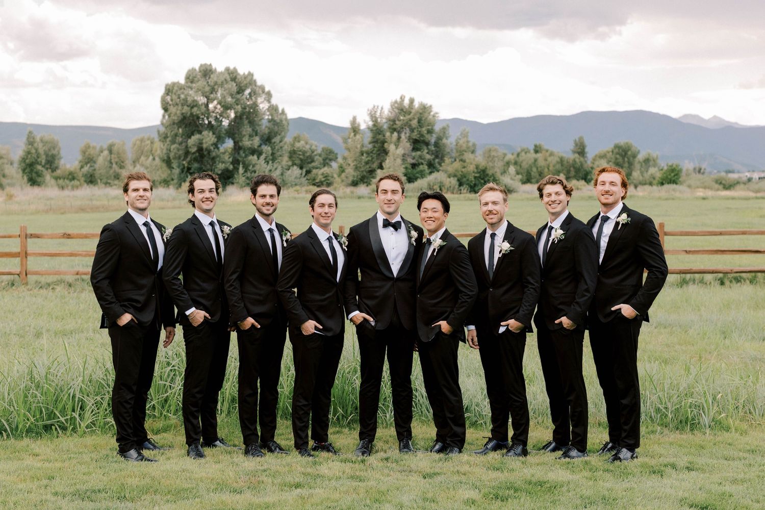 Groomsmen in black tuxedos pose together on rustic ranch with mountains in background.