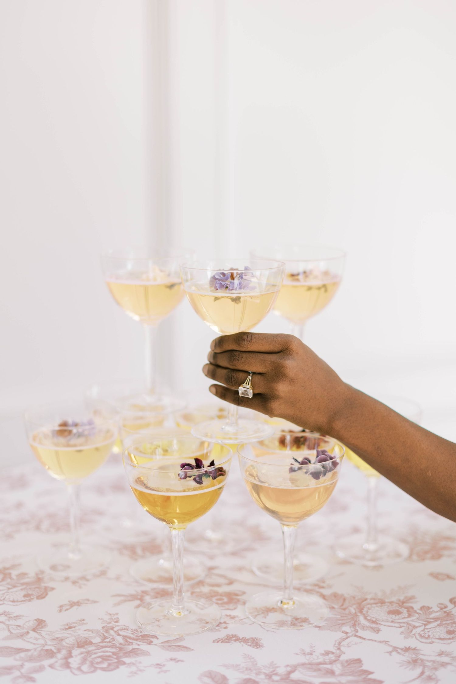 Elegant champagne cocktails garnished with purple flowers are held by a hand wearing an engagement ring.