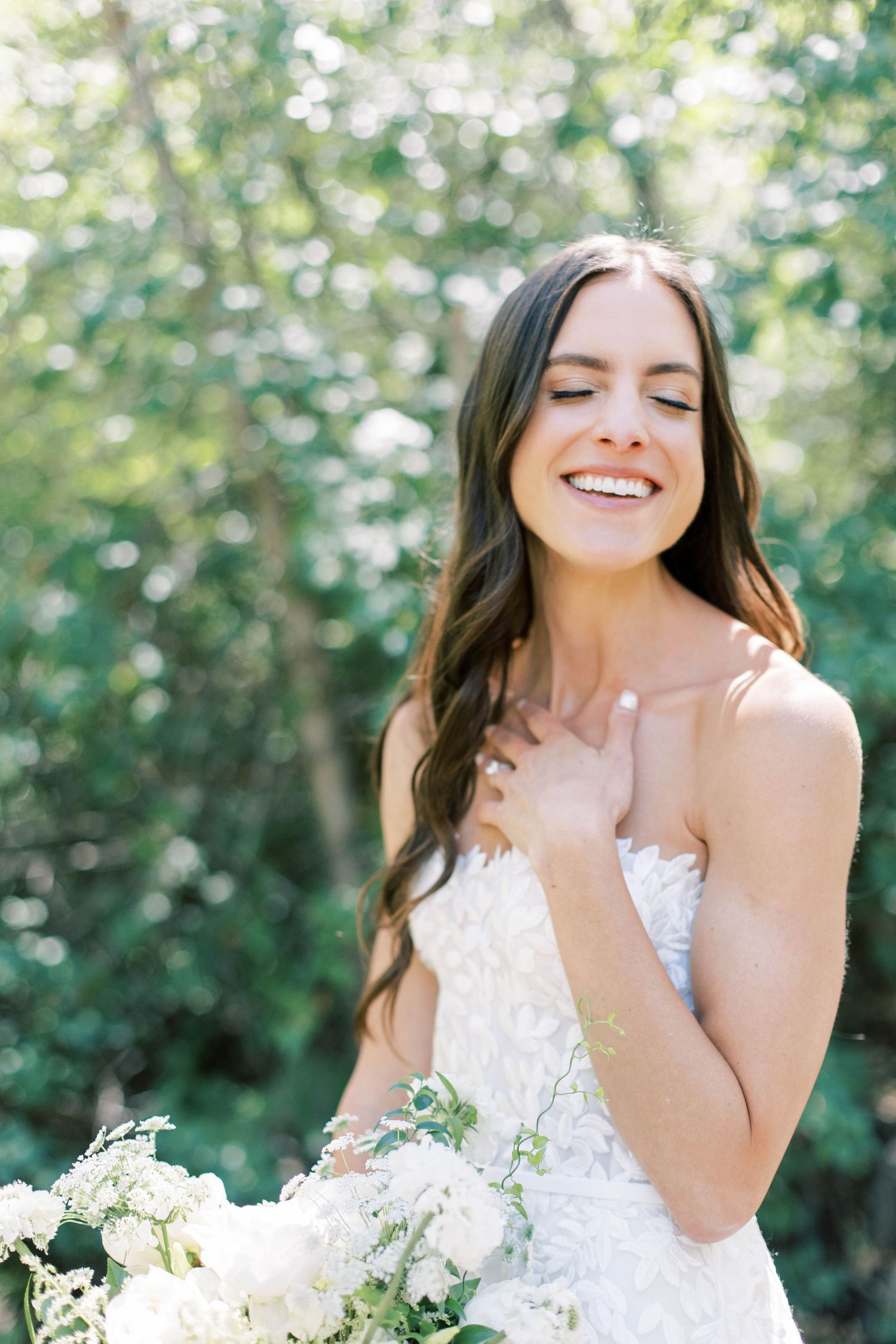 Joyful bride in strapless lace gown holding white bouquet outdoors.