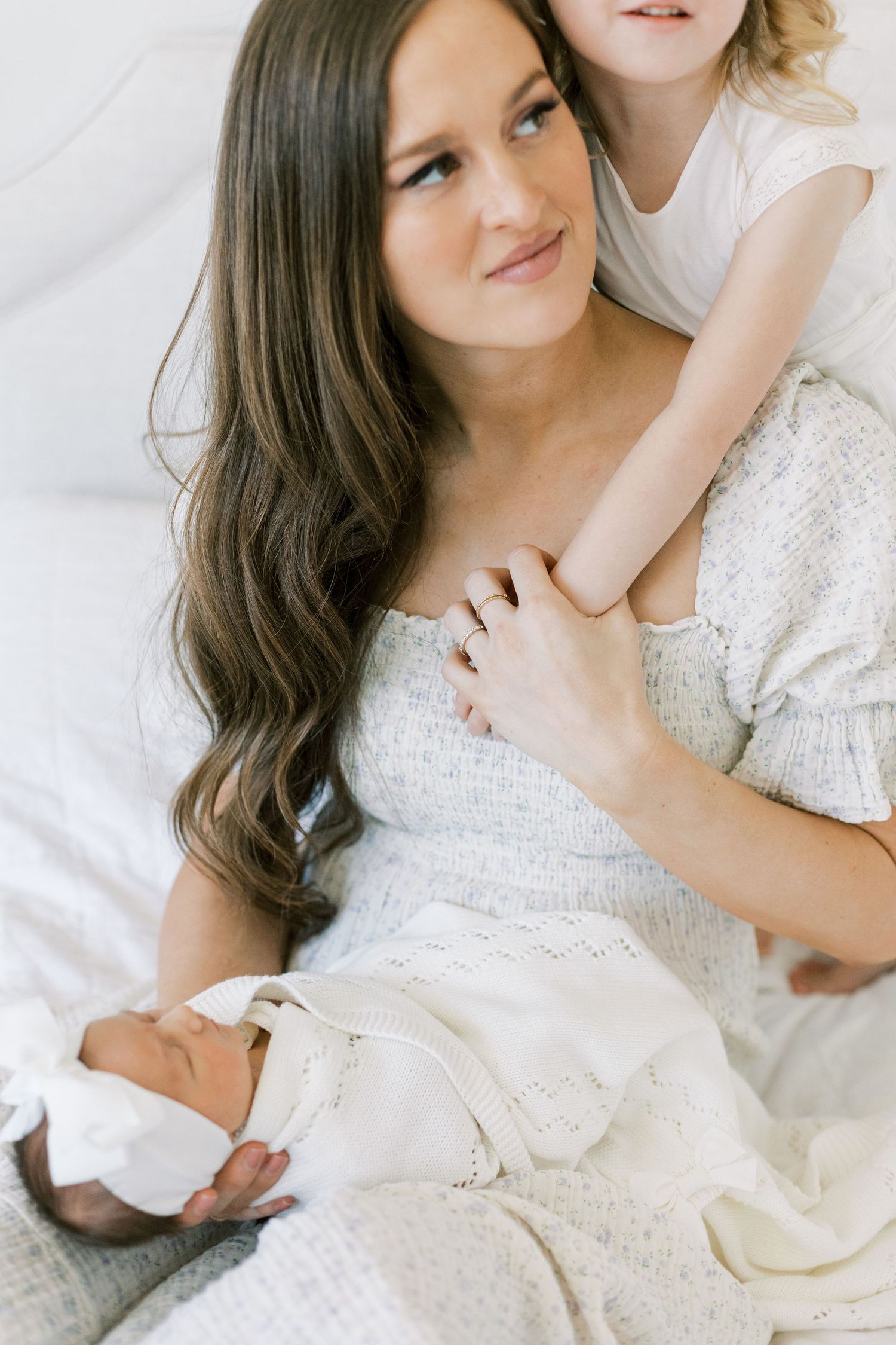 Mother and child in white clothing embrace while sitting on a bed with soft lighting.