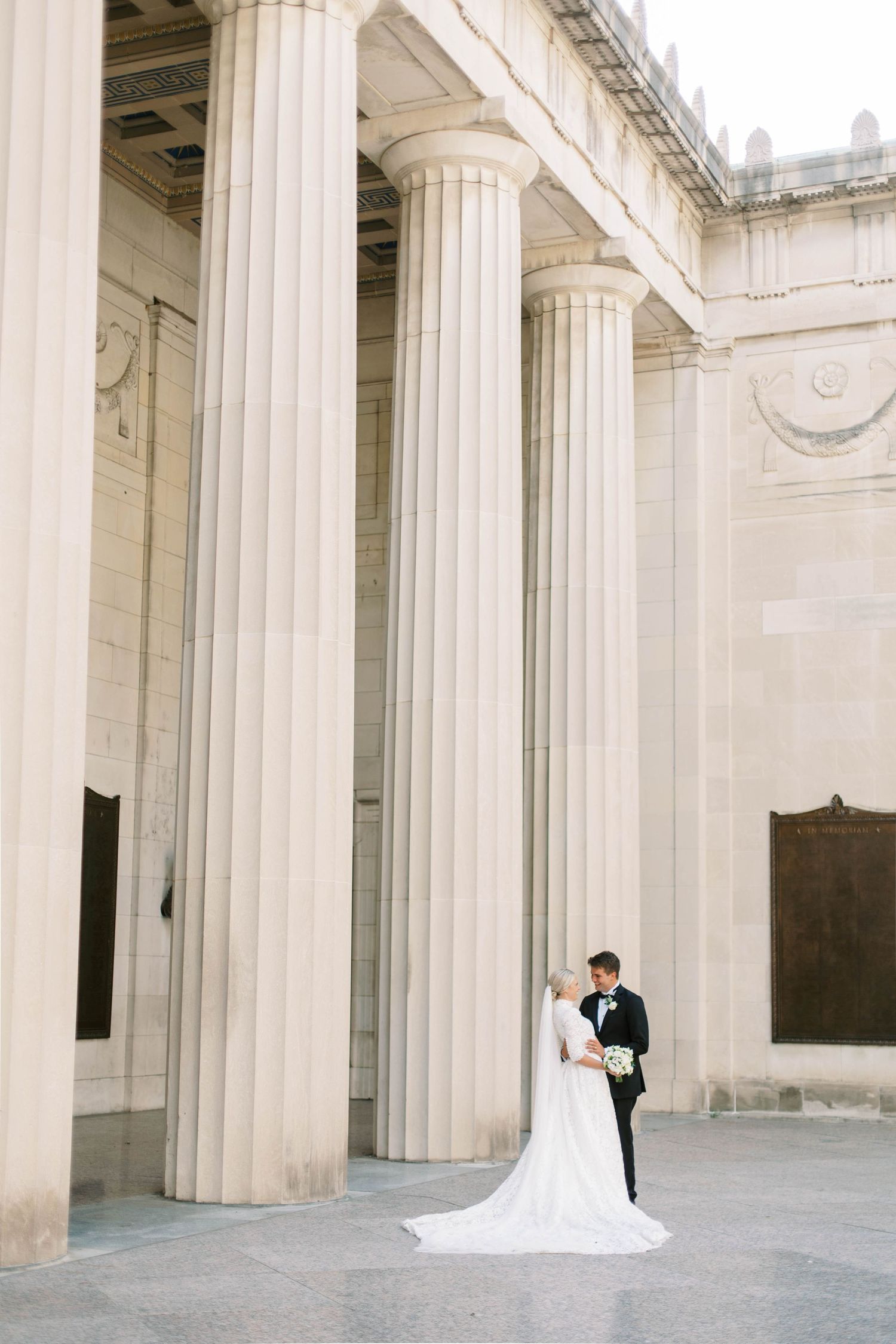 Couple poses against classical white columns in elegant wedding attire.