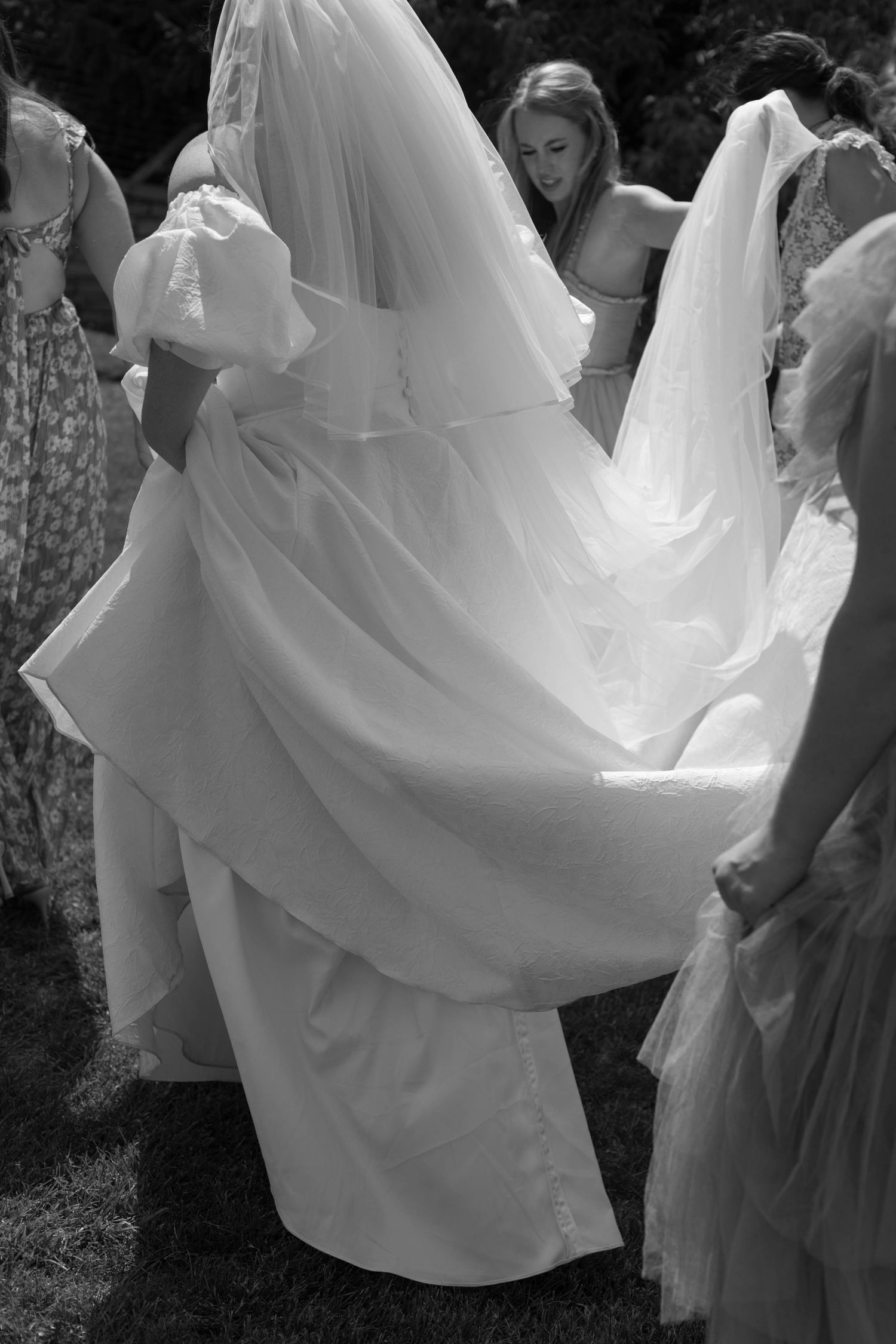Black and white photo of attendants helping with bride's flowing dress train outdoors.
