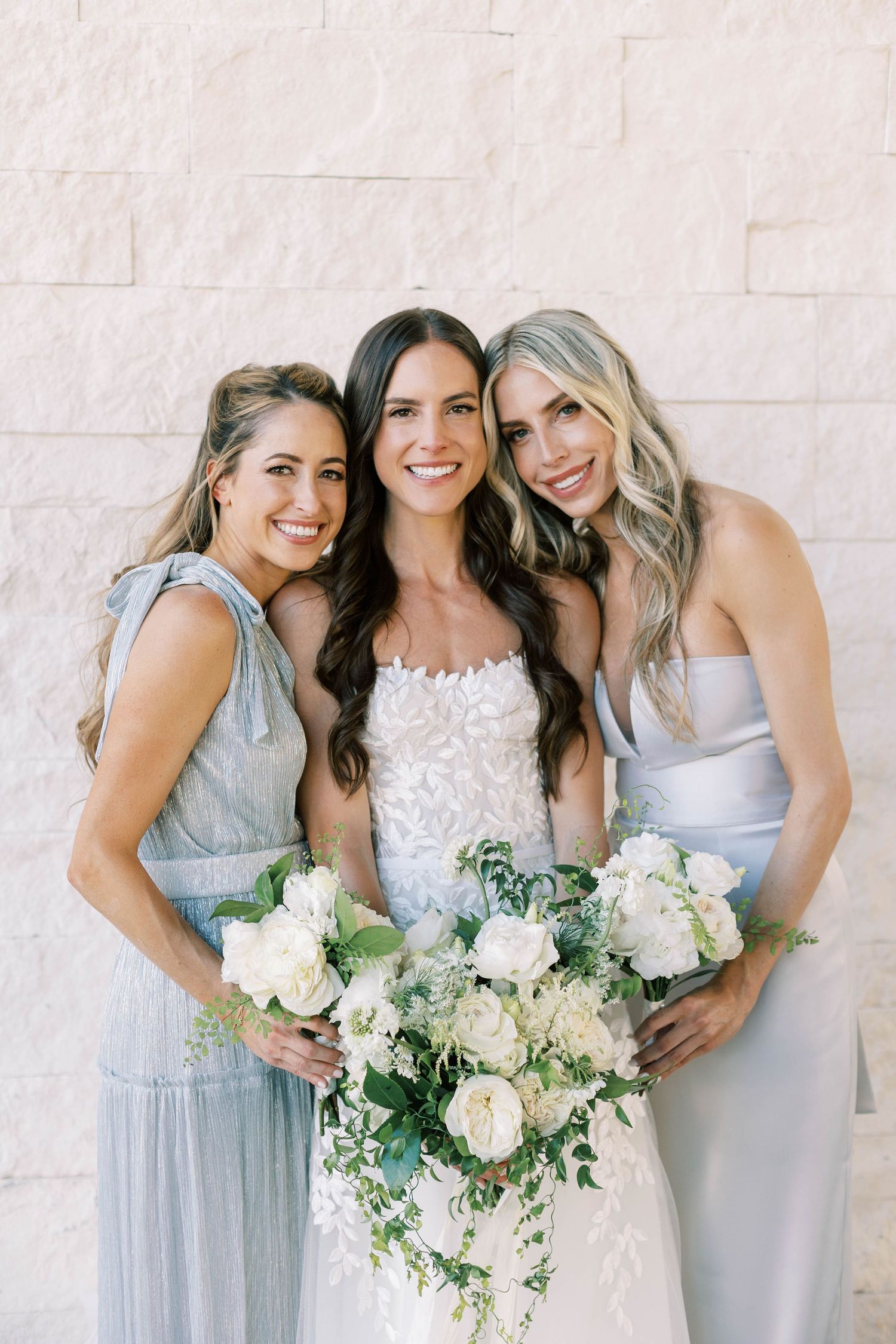 Three women in coordinating wedding attire share smiles while holding white and green floral bouquets.