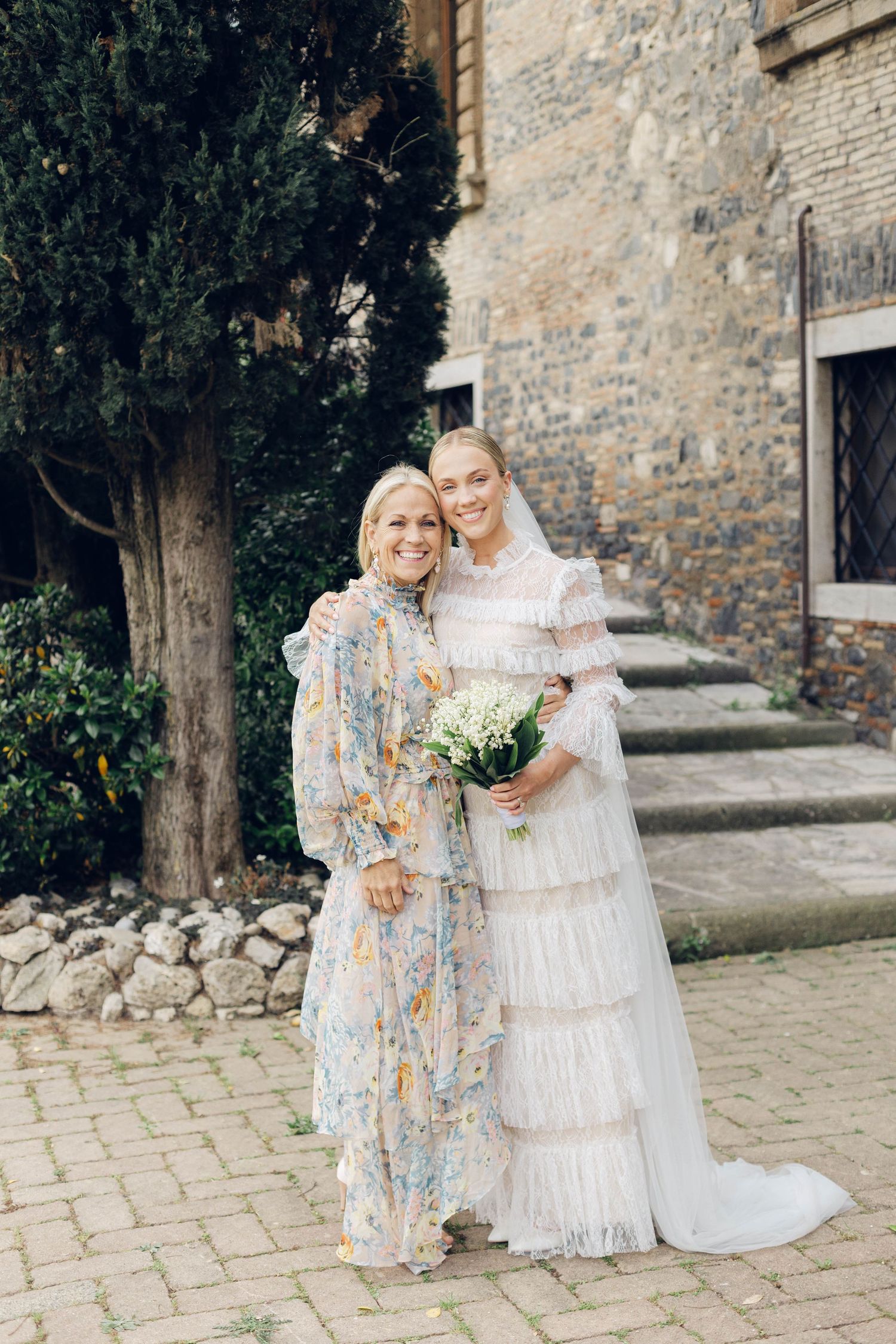 Two wedding guests in elegant attire pose on cobblestone path by stone building.