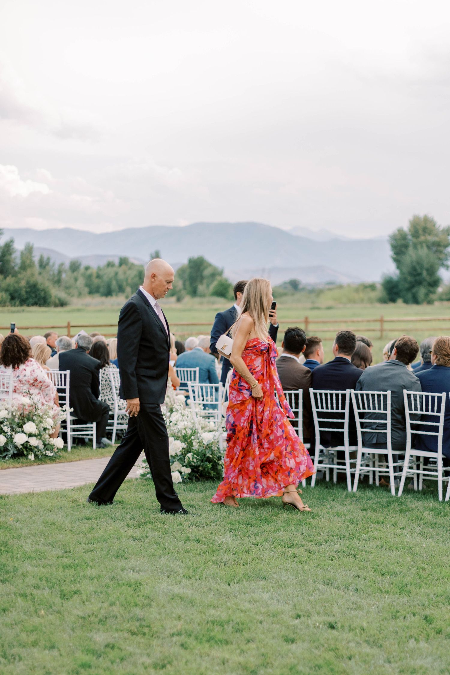Wedding guests walking past white ceremony chairs with mountain backdrop.