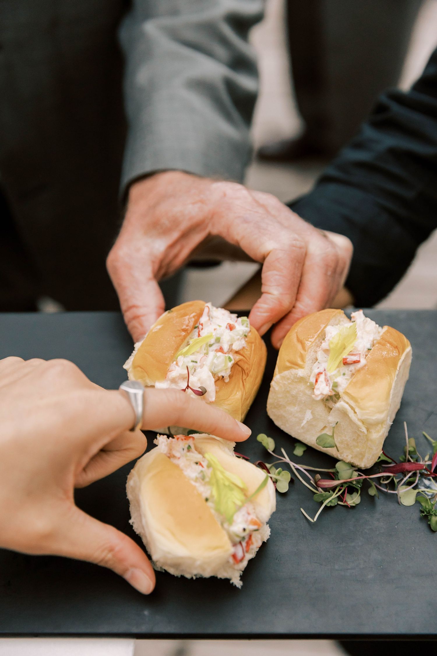 Hands serving small sandwich sliders garnished with microgreens on a dark surface.