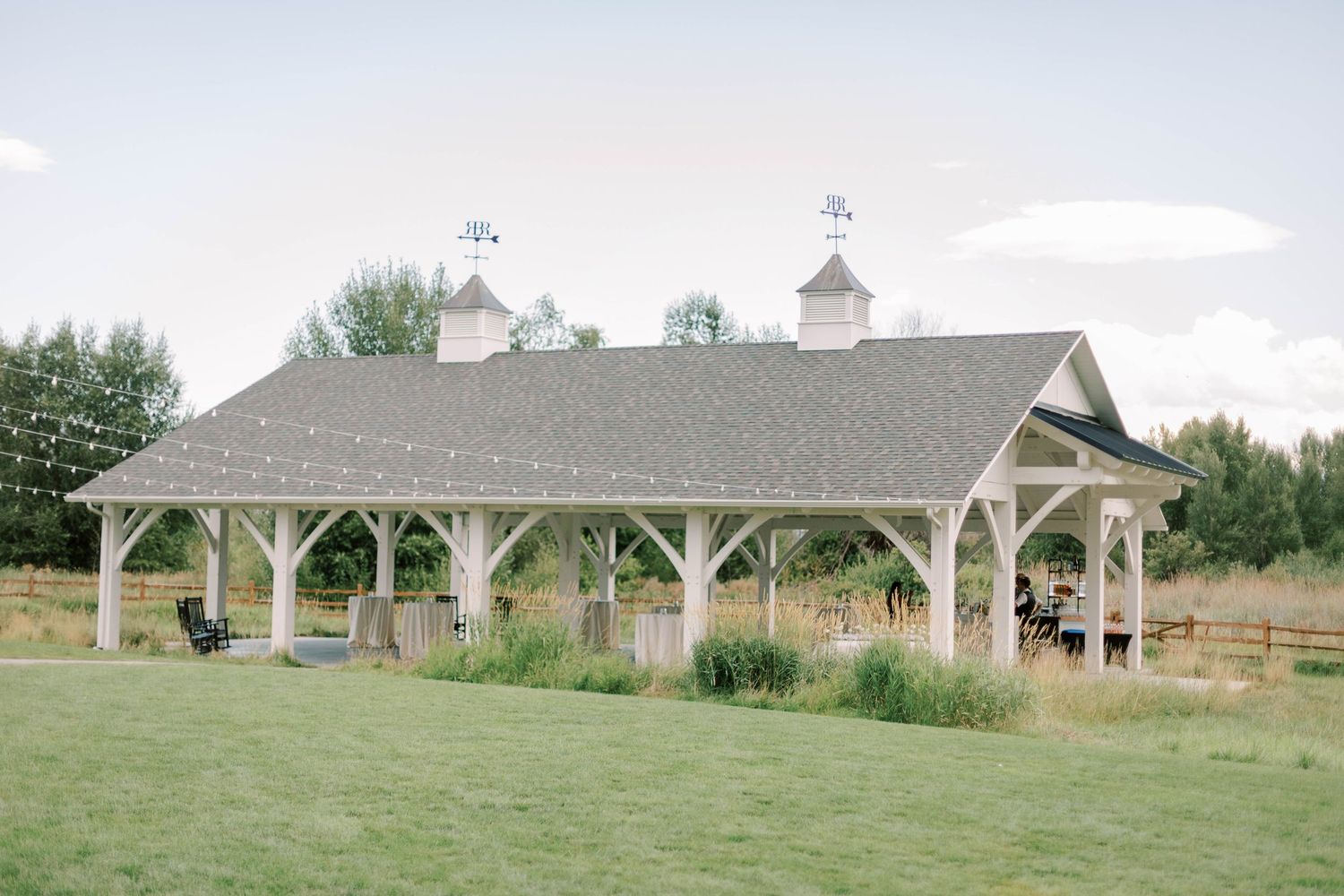 A rustic white outdoor pavilion with wooden posts and gable roof set in a grassy field.