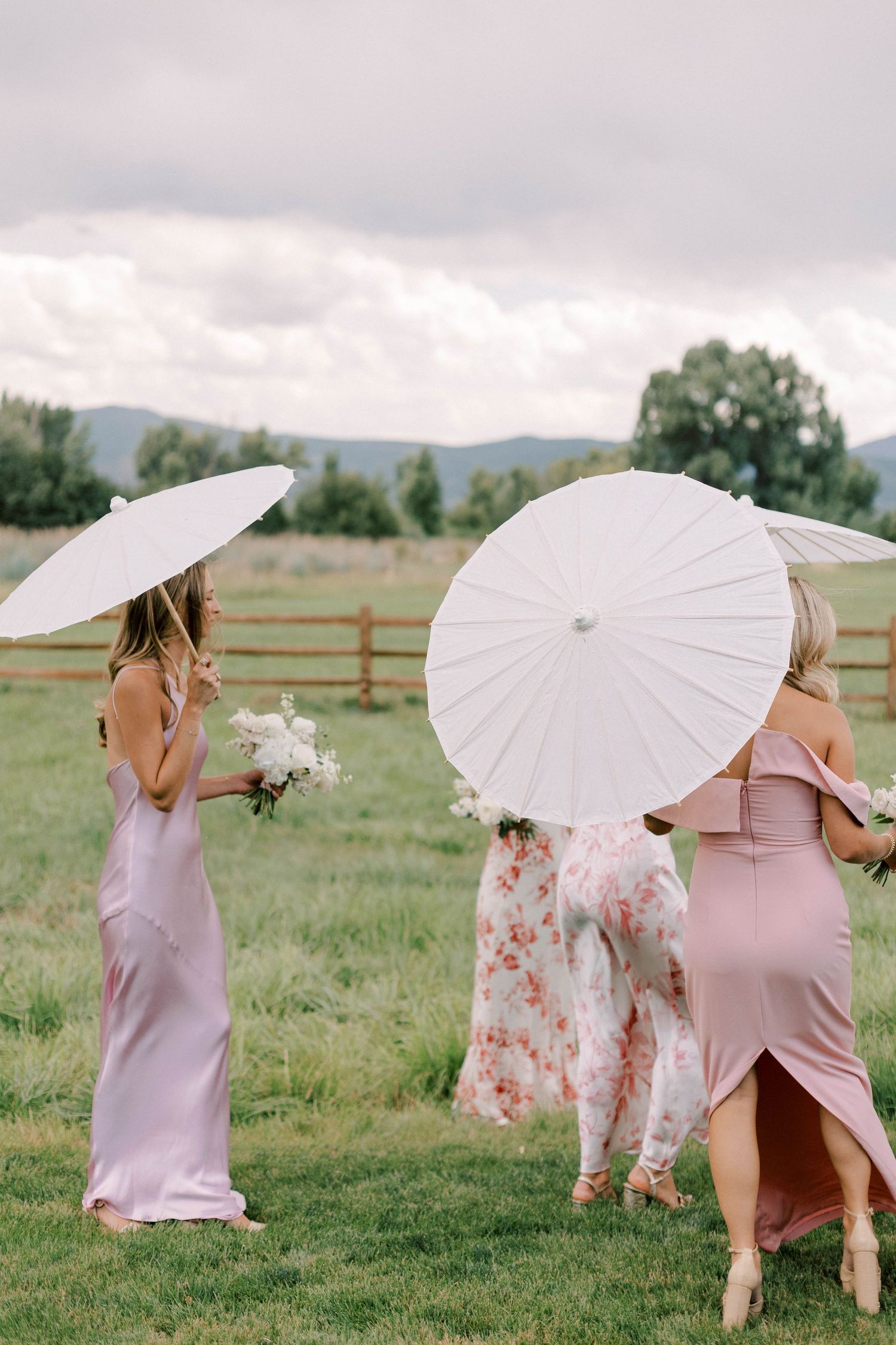 Bridesmaids in pink dresses holding white parasols on a grassy field.