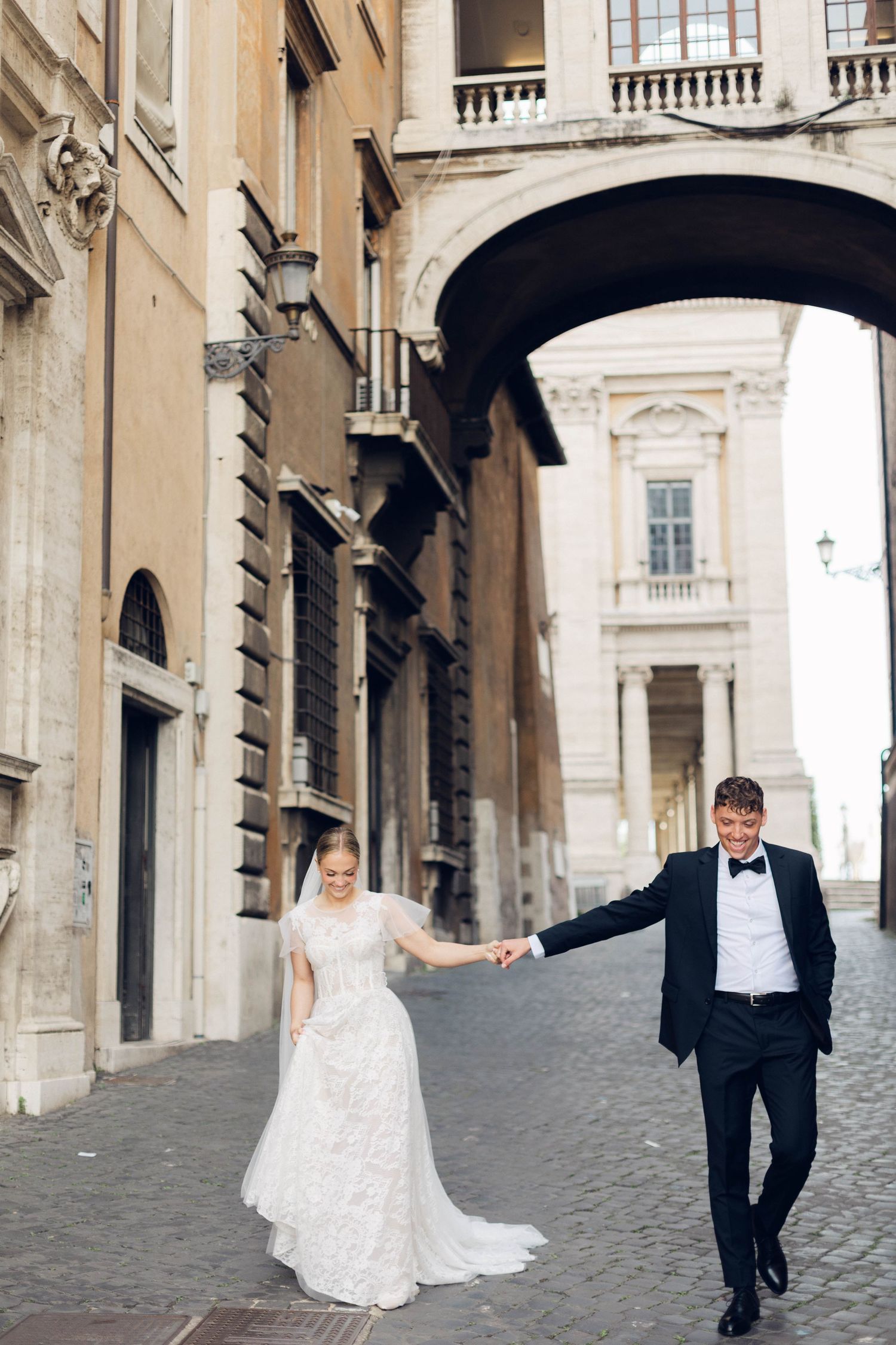 Joyful wedding couple holding hands while walking through historic Roman street passage.