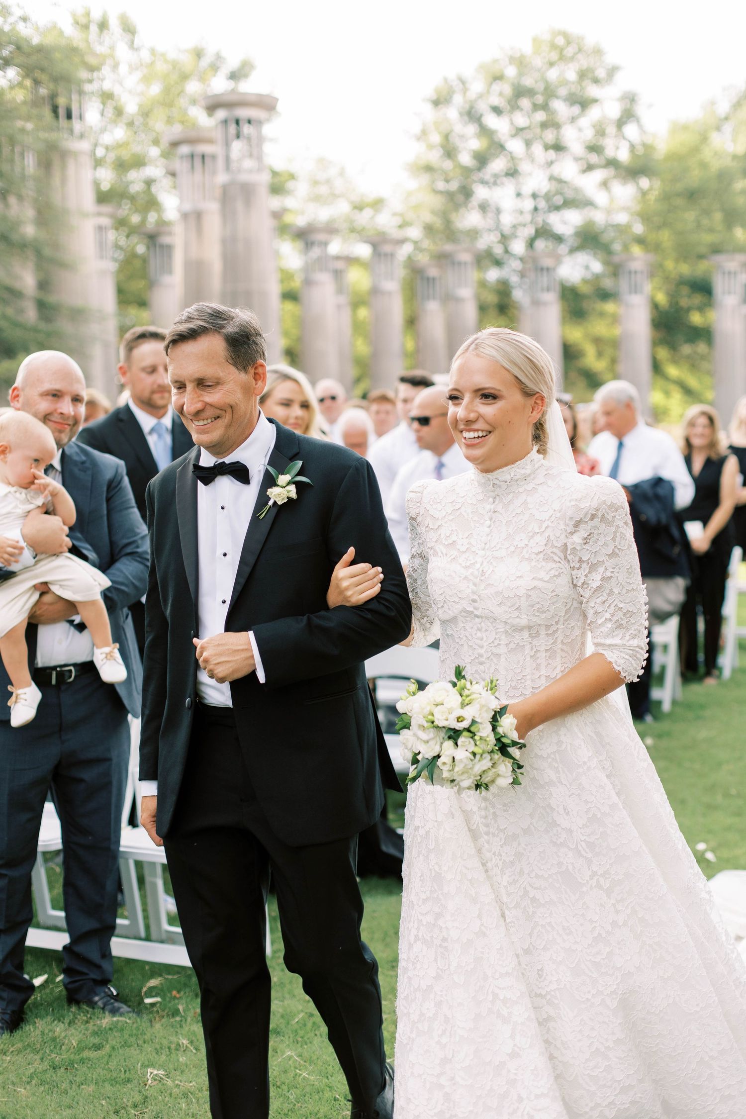 Wedding ceremony taking place outdoors with stone columns and guests gathered on a lawn.