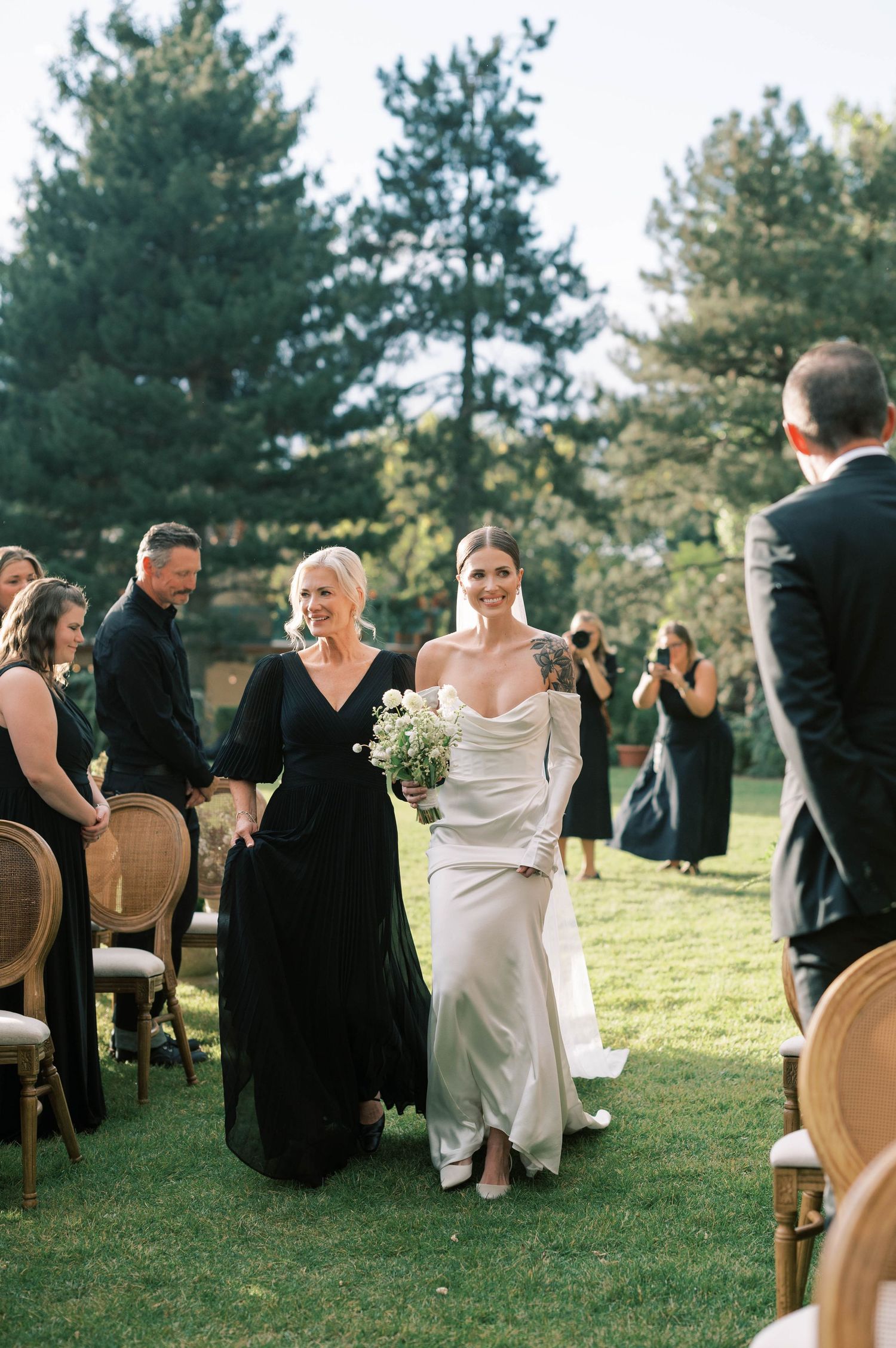 Wedding ceremony procession with attendees in formal black attire on manicured lawn.