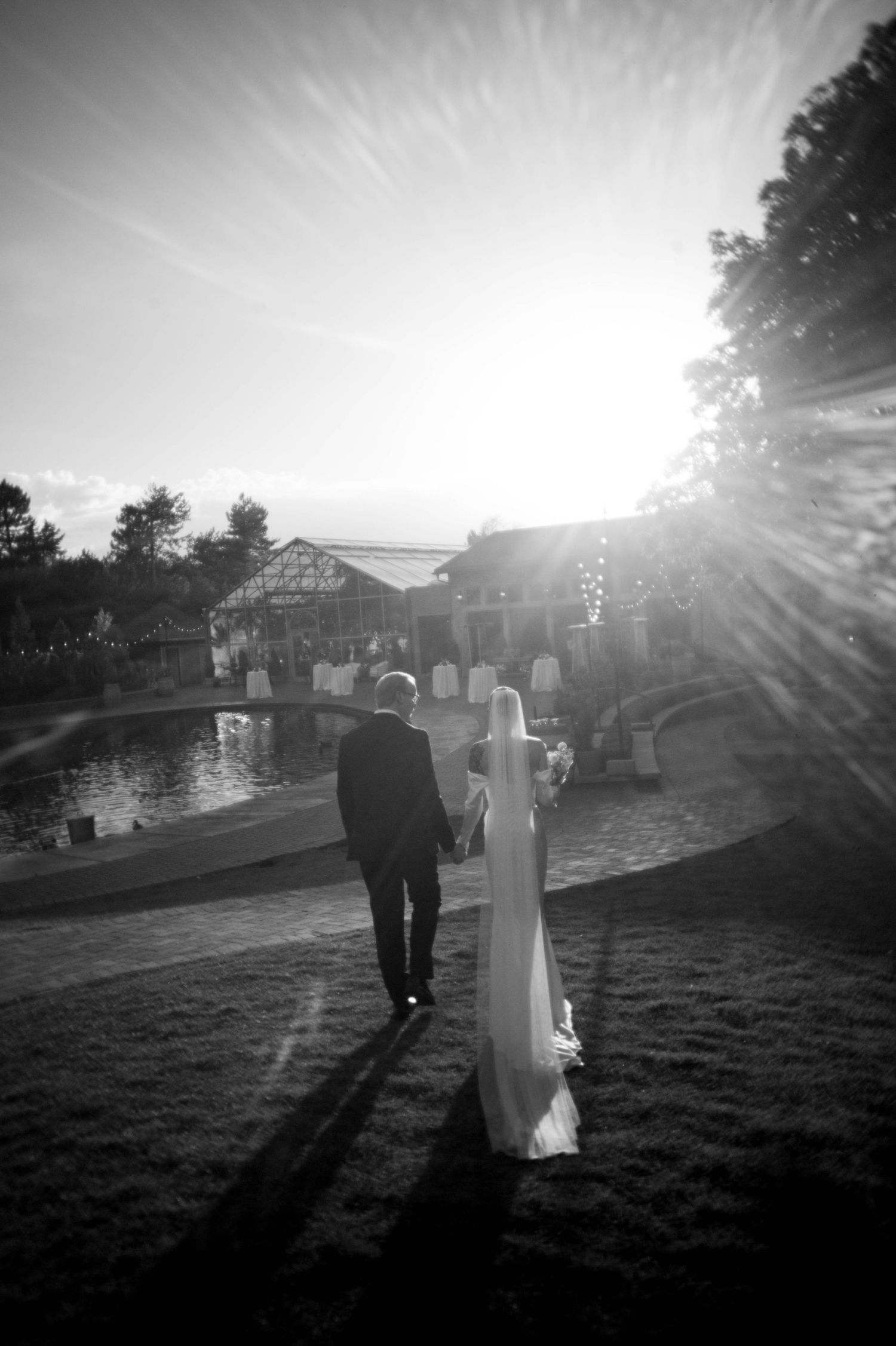 A black and white silhouette of a wedding couple walking at sunset near a waterfront venue.