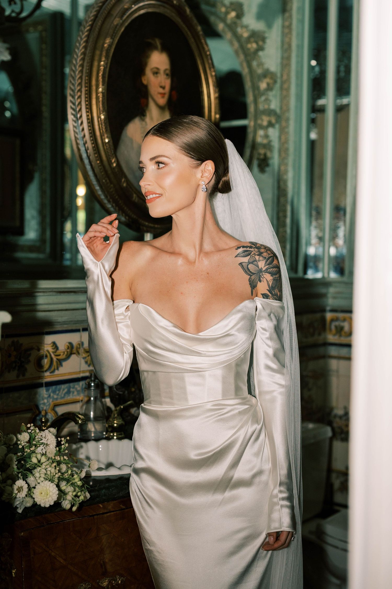 Elegant bride in silk off-shoulder gown with floral tattoo poses in ornate mirror room.