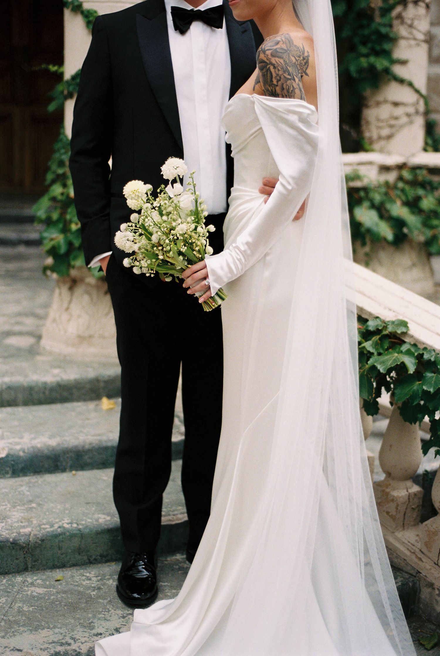 Formal wedding couple in black tuxedo and white gown holding delicate floral bouquet.