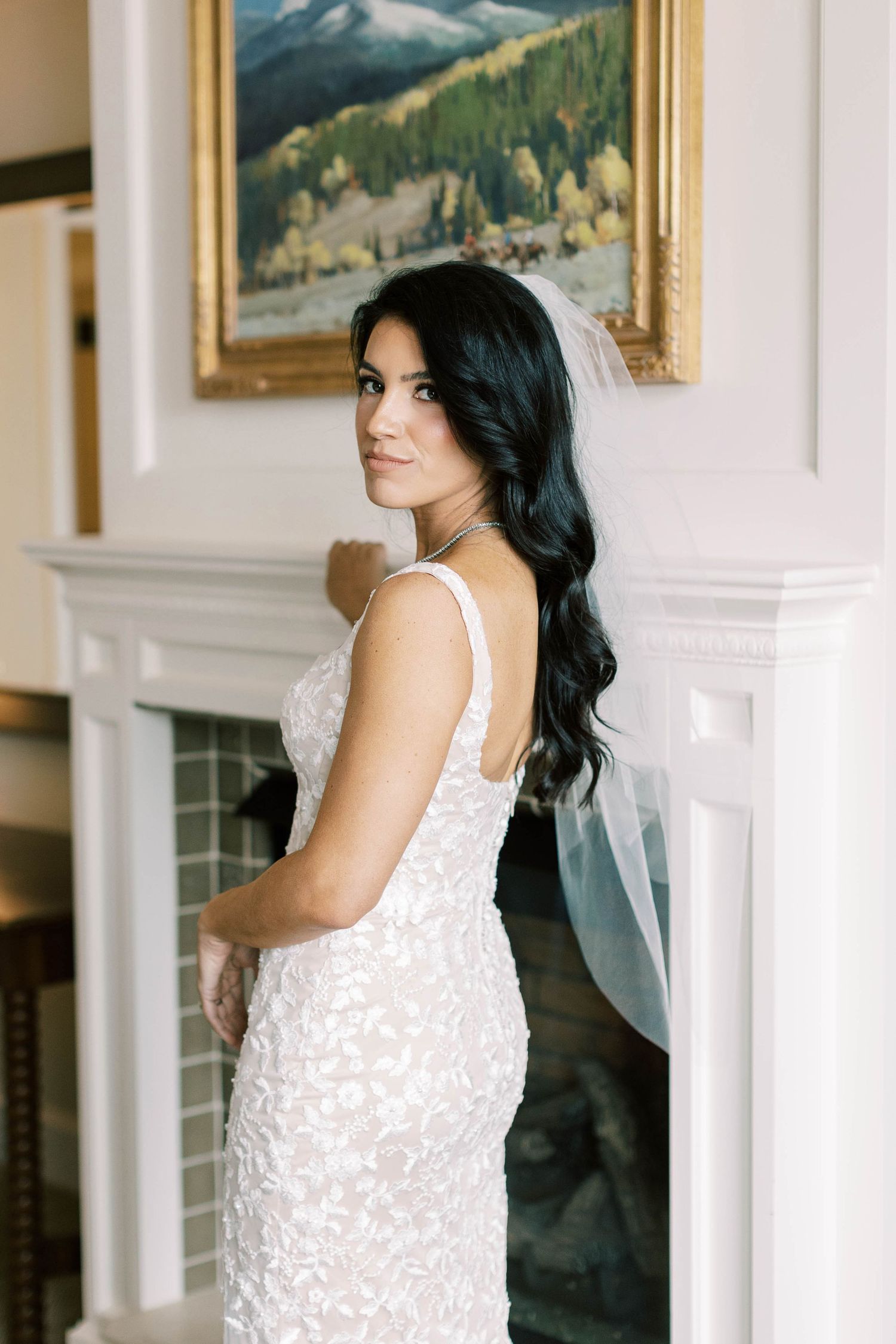 Wedding dress detail with lace pattern and veil against white fireplace mantel and mountain artwork.