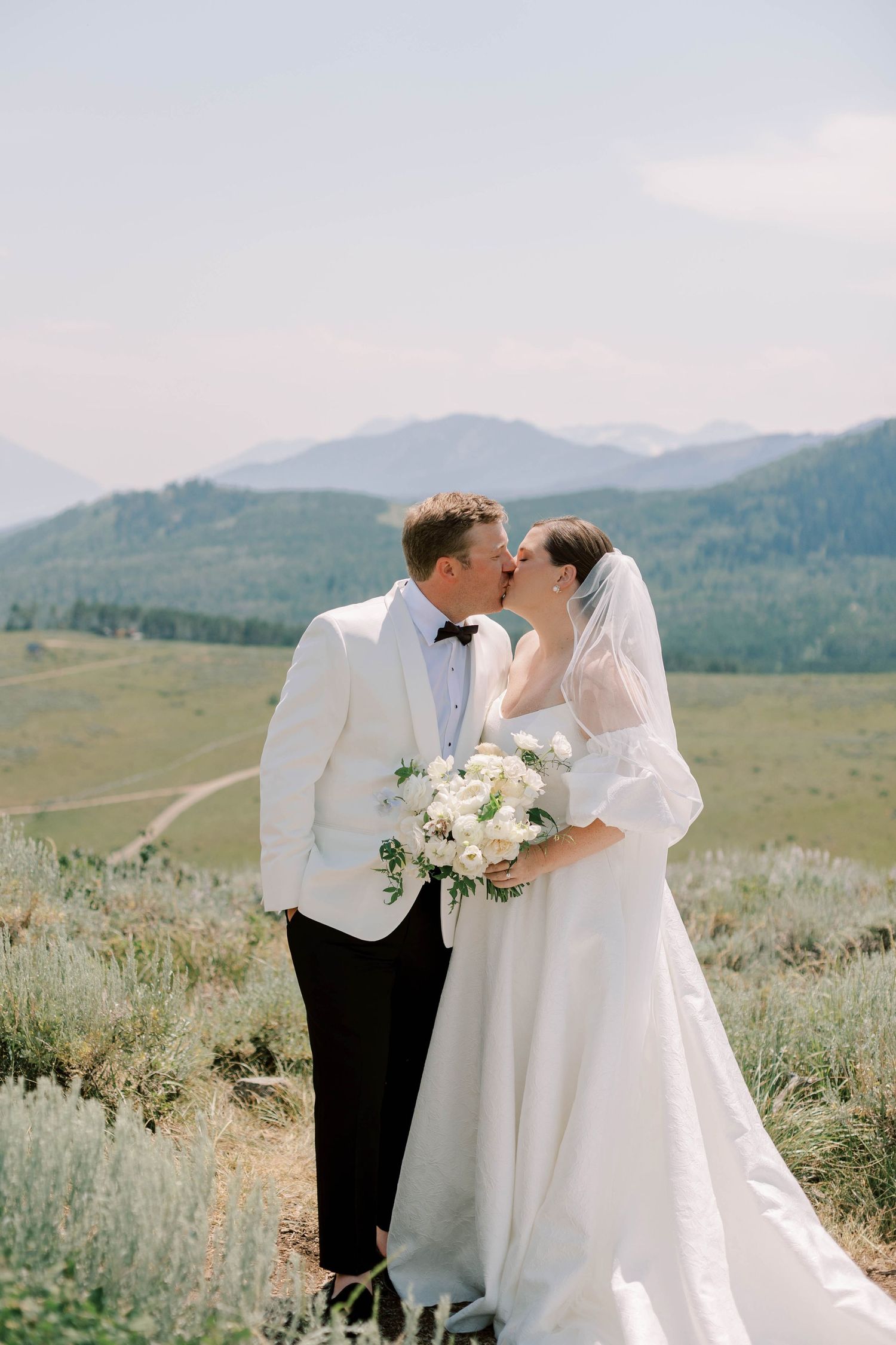Newlyweds kiss while holding white bouquet against mountain backdrop.