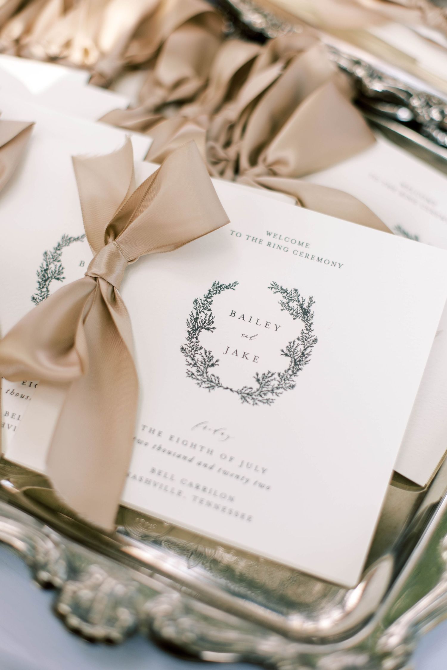 Elegant wedding invitation with botanical wreath design and champagne colored ribbon on silver tray.