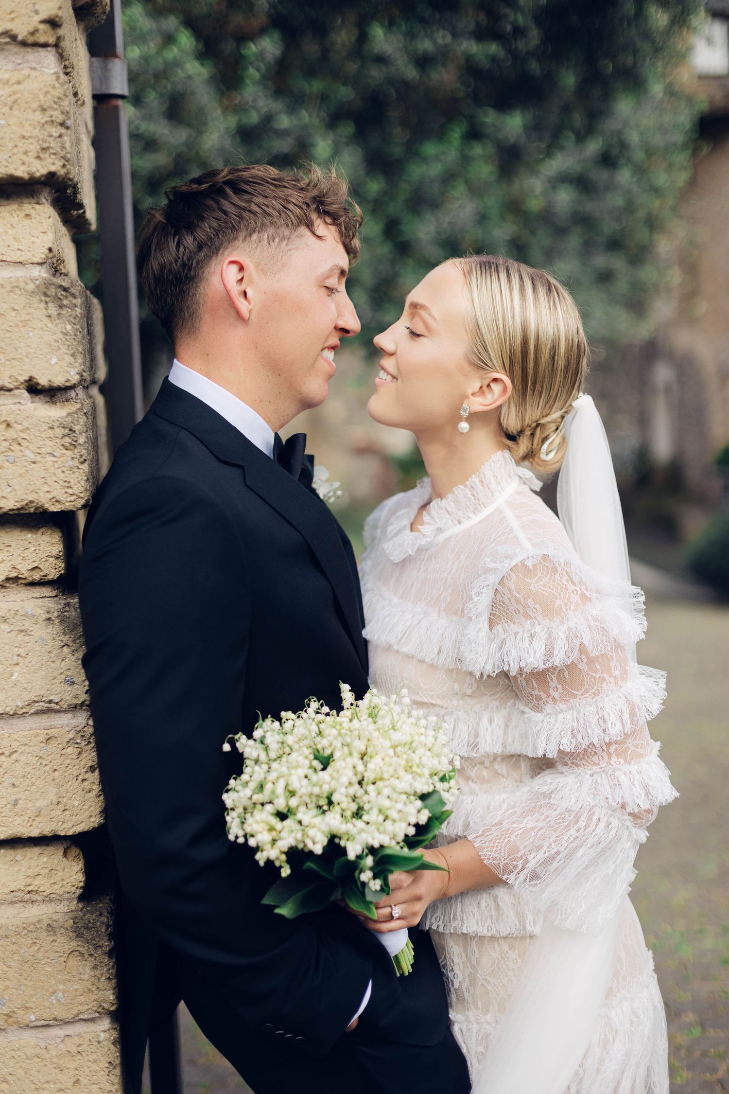 Romantic moment captured between newlyweds against a stone wall with a bouquet of white flowers.