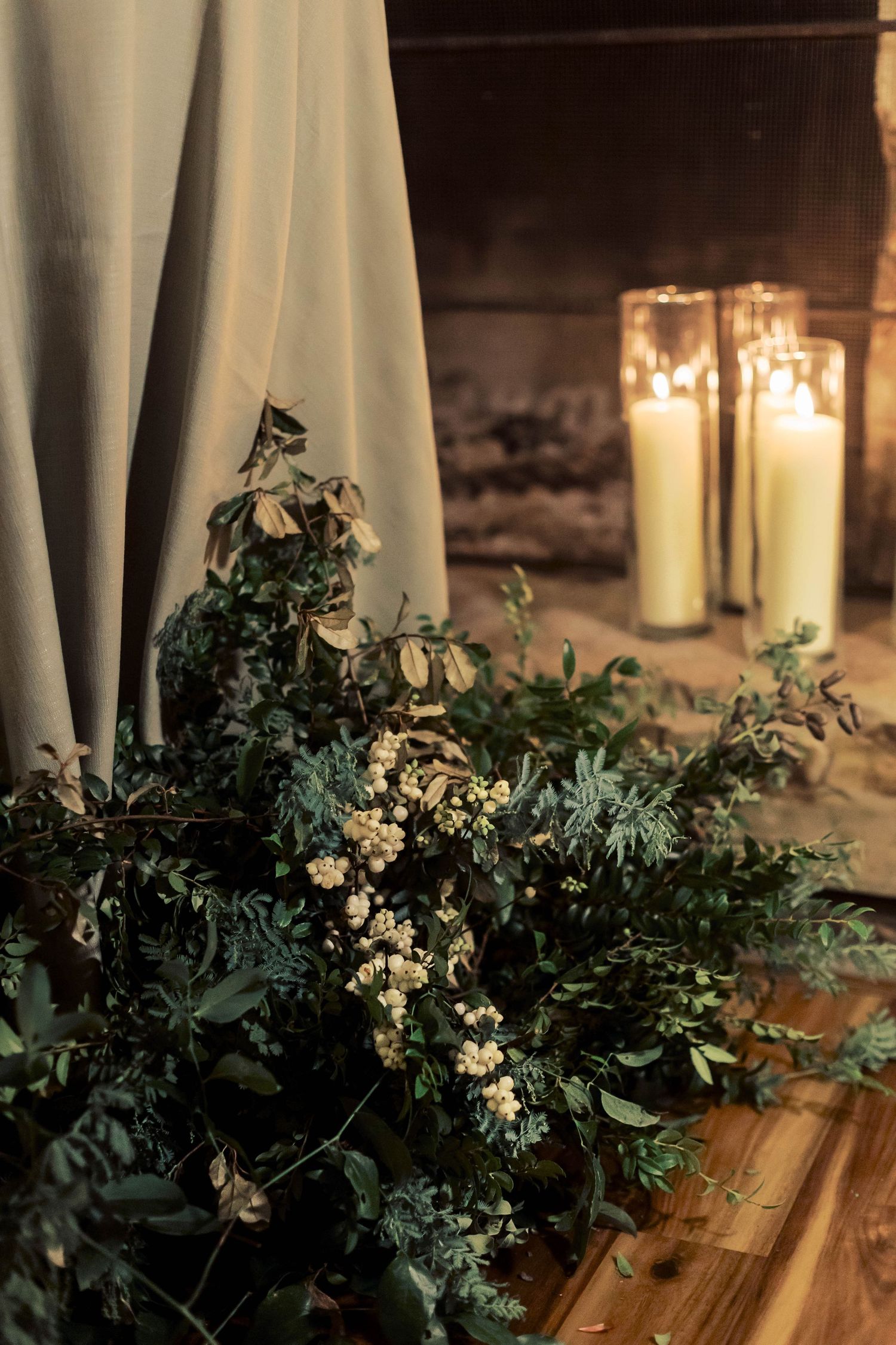 Moody floral arrangement with white flowers and greenery next to glowing candles against draped fabric.