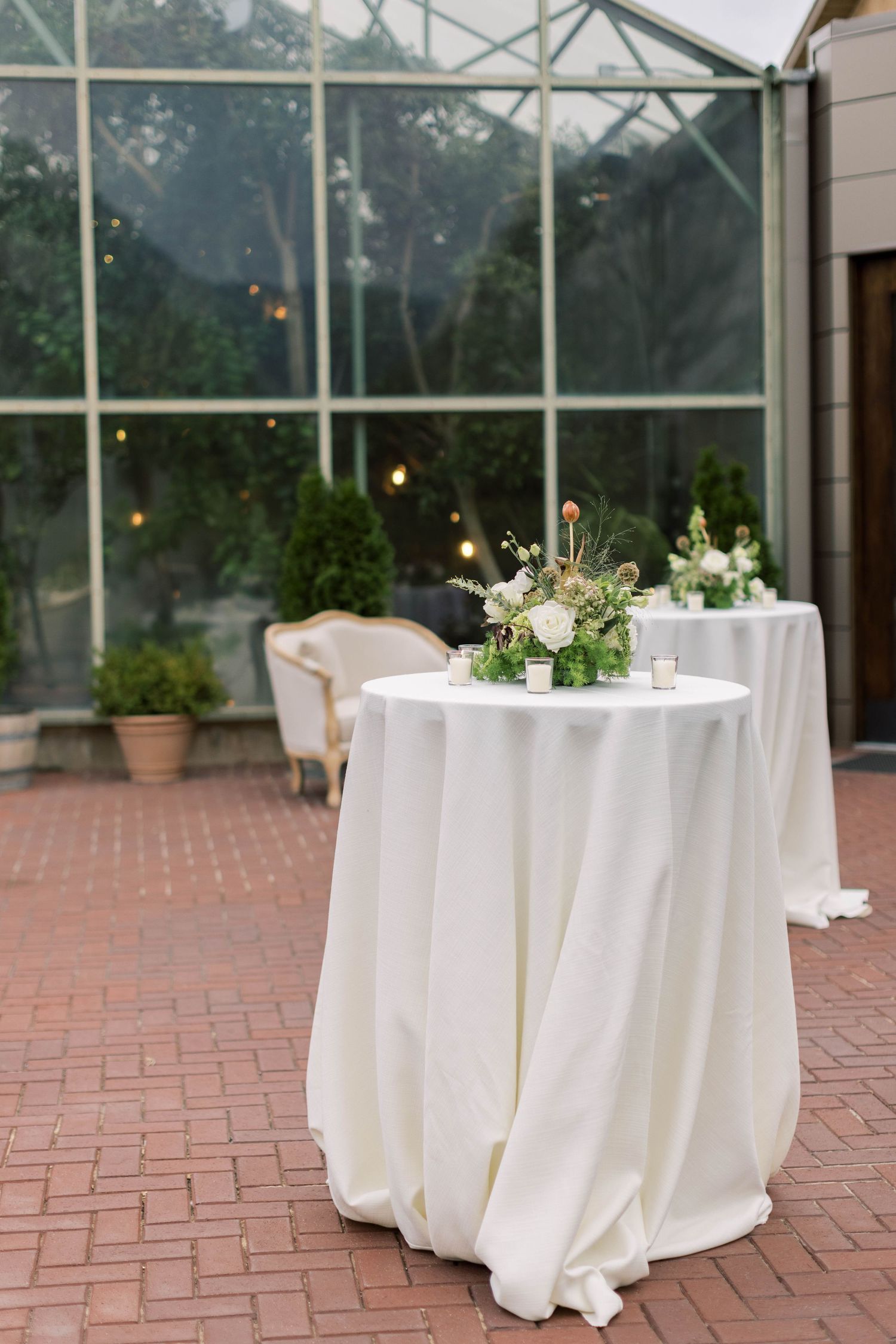 White cocktail table with floral arrangement against glass greenhouse backdrop.
