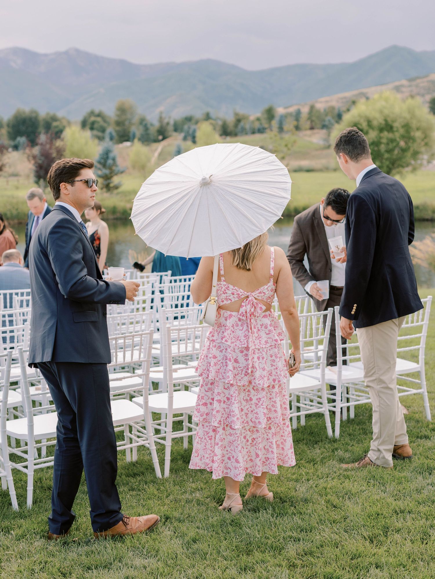 Guests gathering at outdoor wedding ceremony with mountains and parasol.