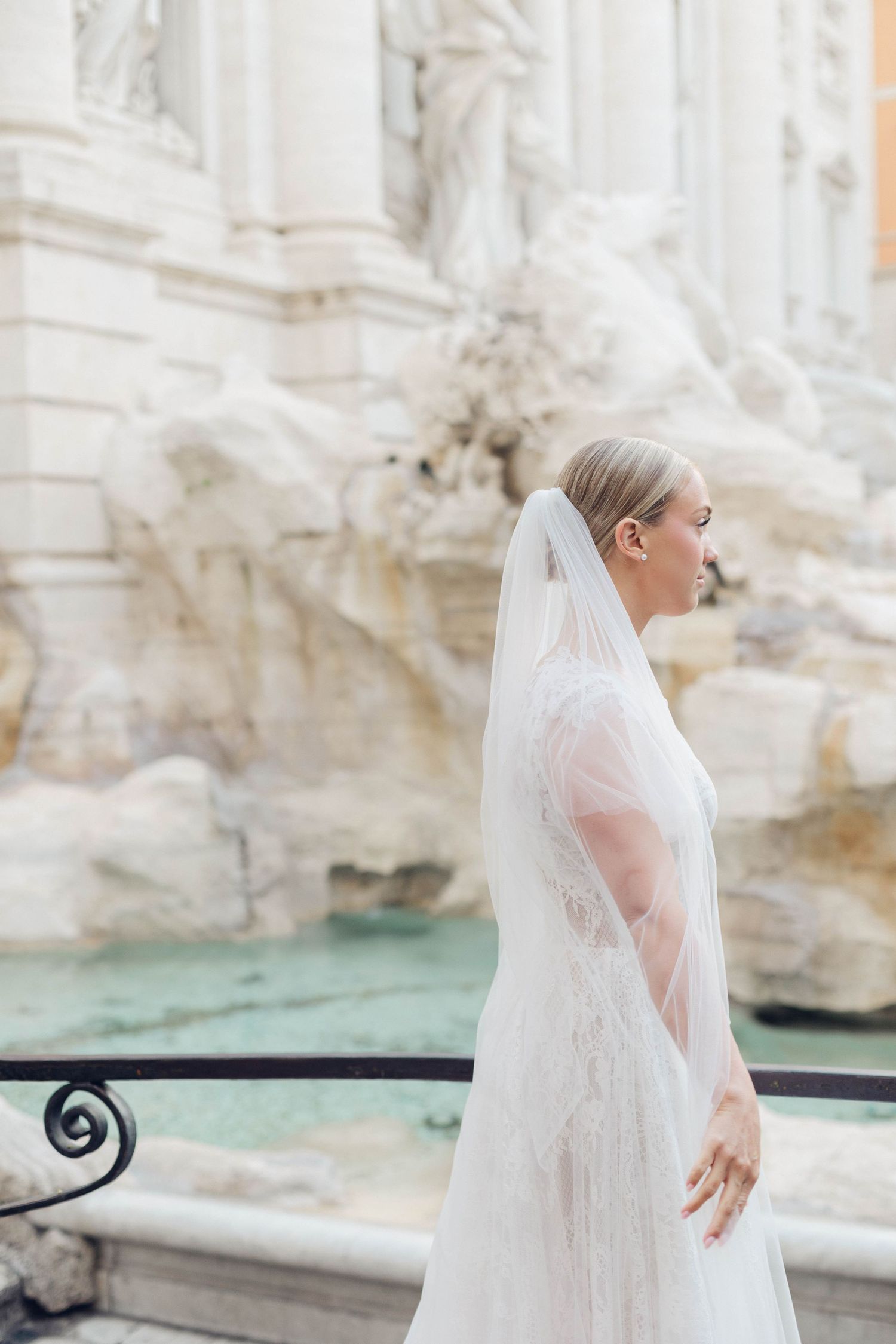 Side profile of bride in delicate lace gown standing beside the turquoise waters of Trevi Fountain.