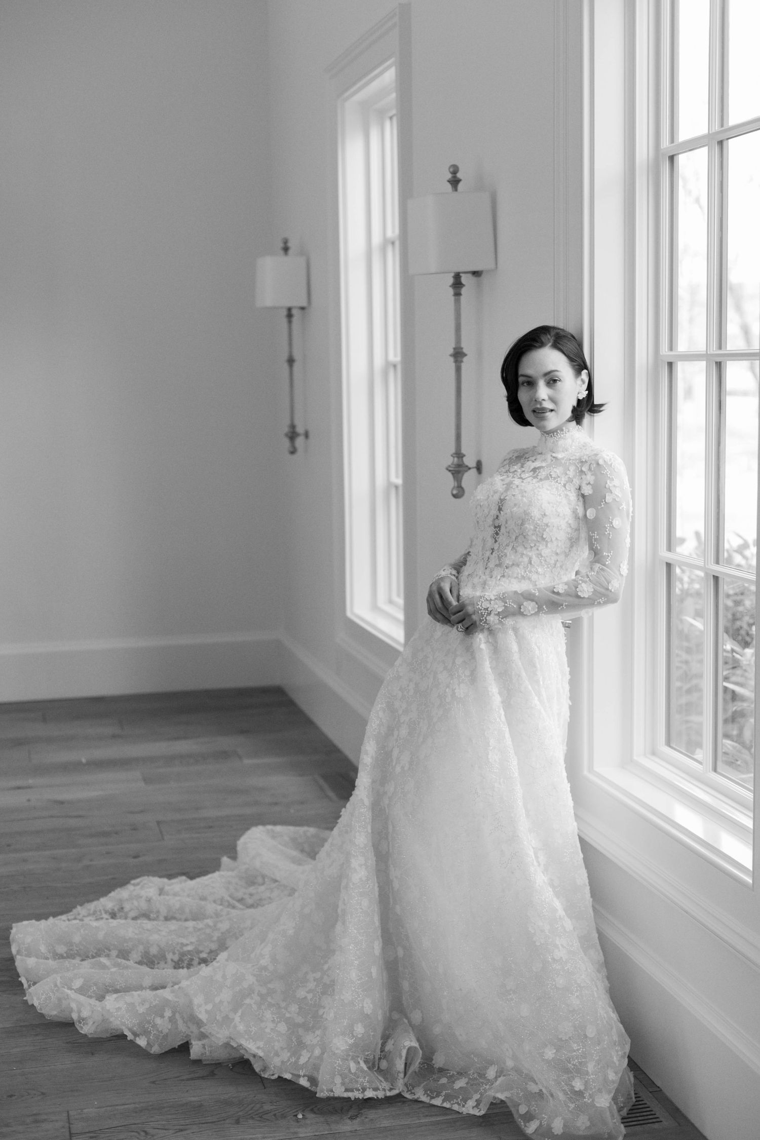 An elegant long-sleeve lace wedding gown photographed in a bright interior setting.