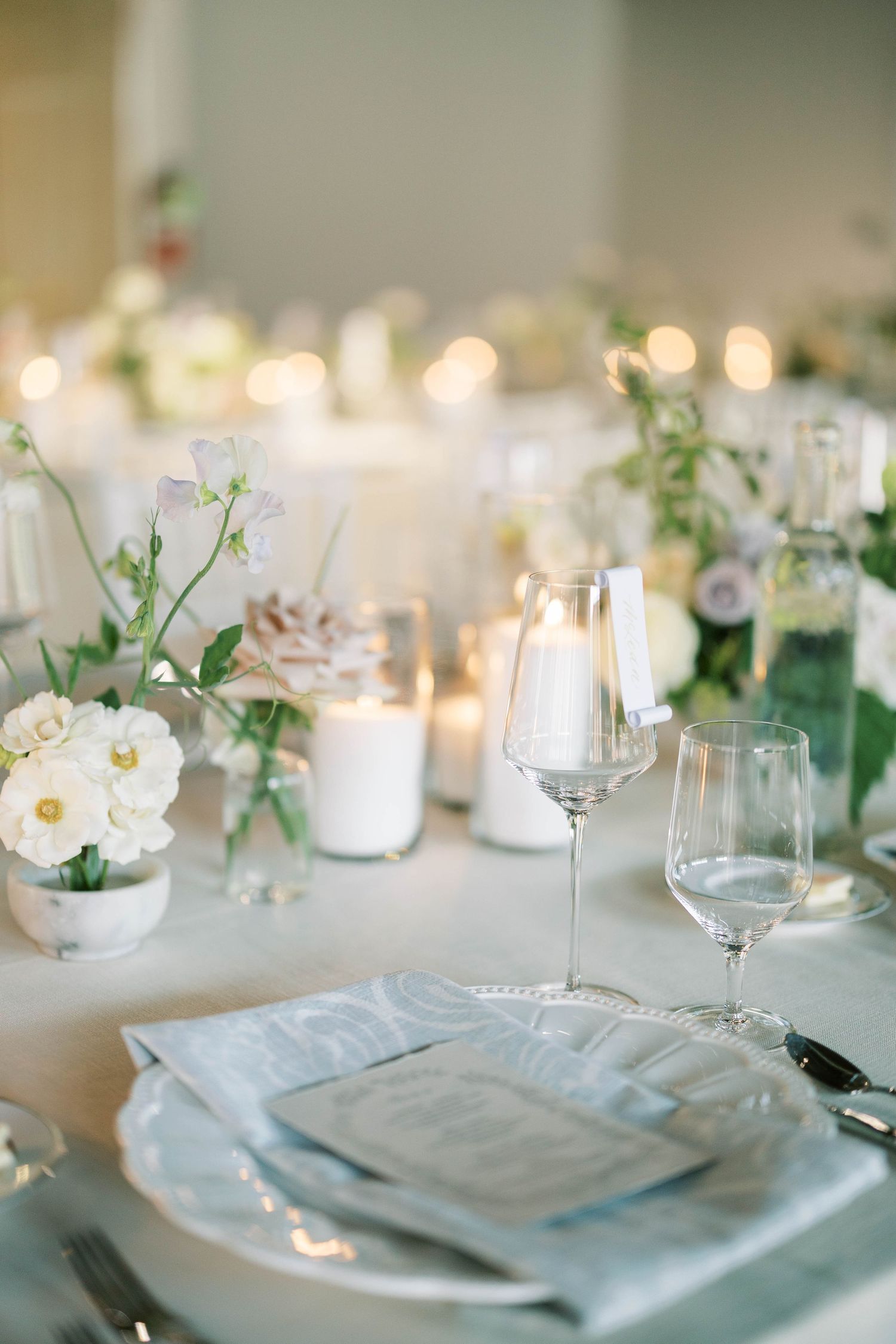 Romantic table setting with white flowers, candles, and wine glasses on light gray tablecloth.