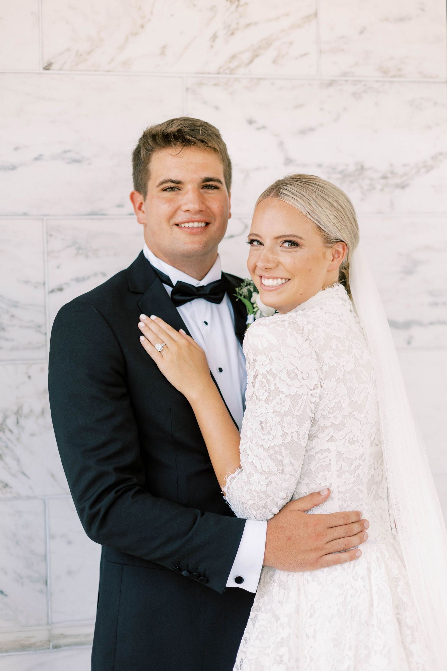 Newlyweds pose together against marble wall, bride in lace dress and groom in tuxedo with boutonniere.