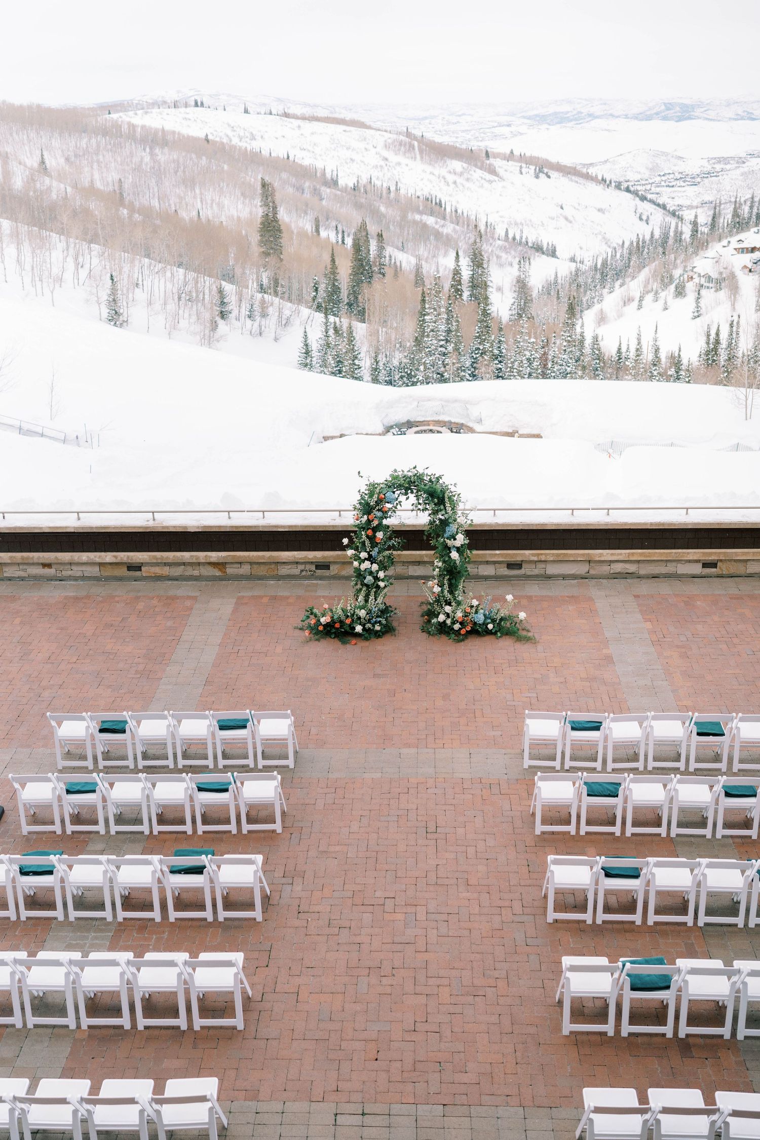 Snow-covered mountain view behind an outdoor wedding ceremony setup with white chairs and floral arch.