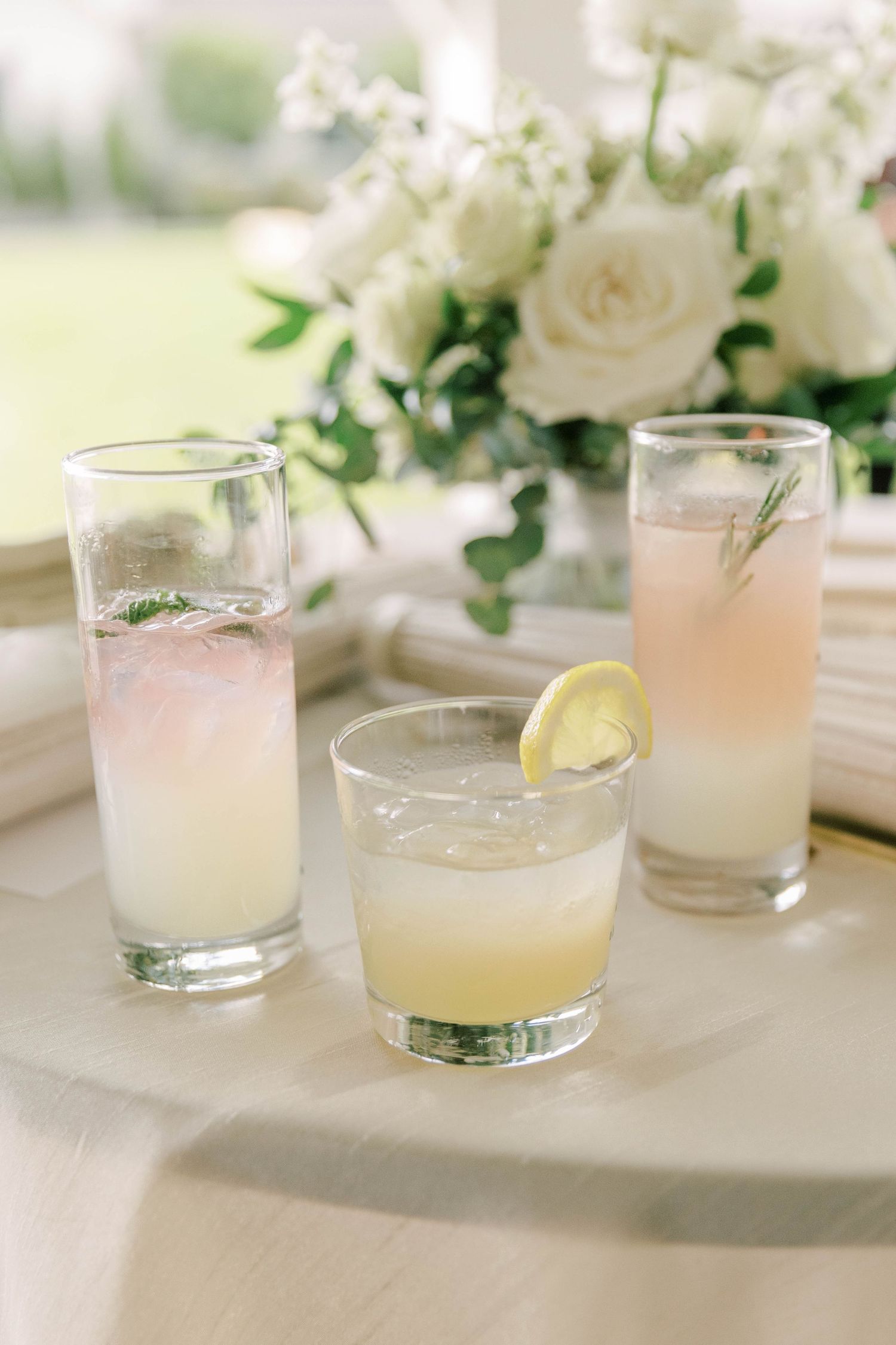 Three light pink cocktails with citrus garnishes and white floral centerpiece on table.