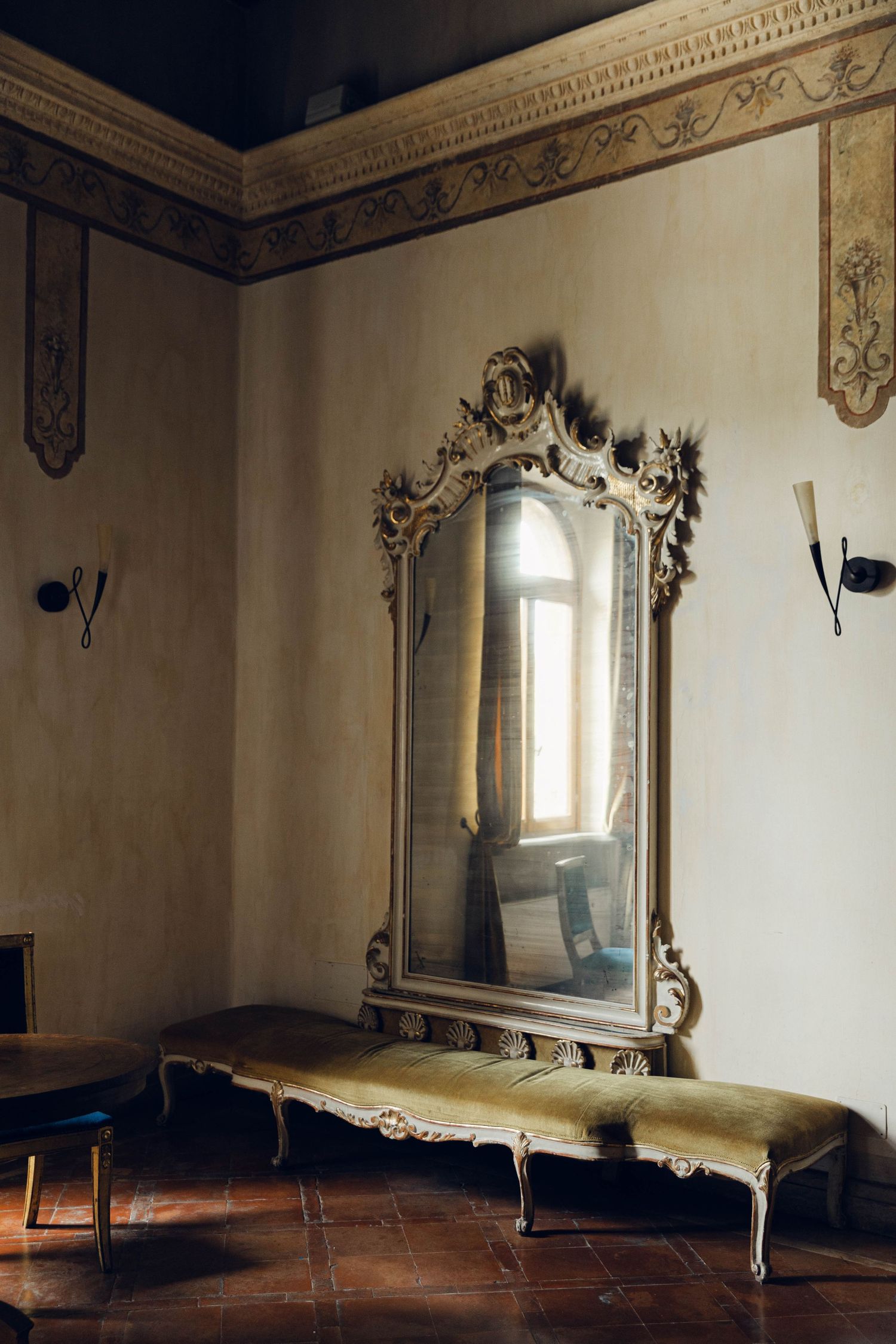 Ornate gilded mirror with baroque frame mounted above antique bench in grand room in castello della castellucia rome italy.