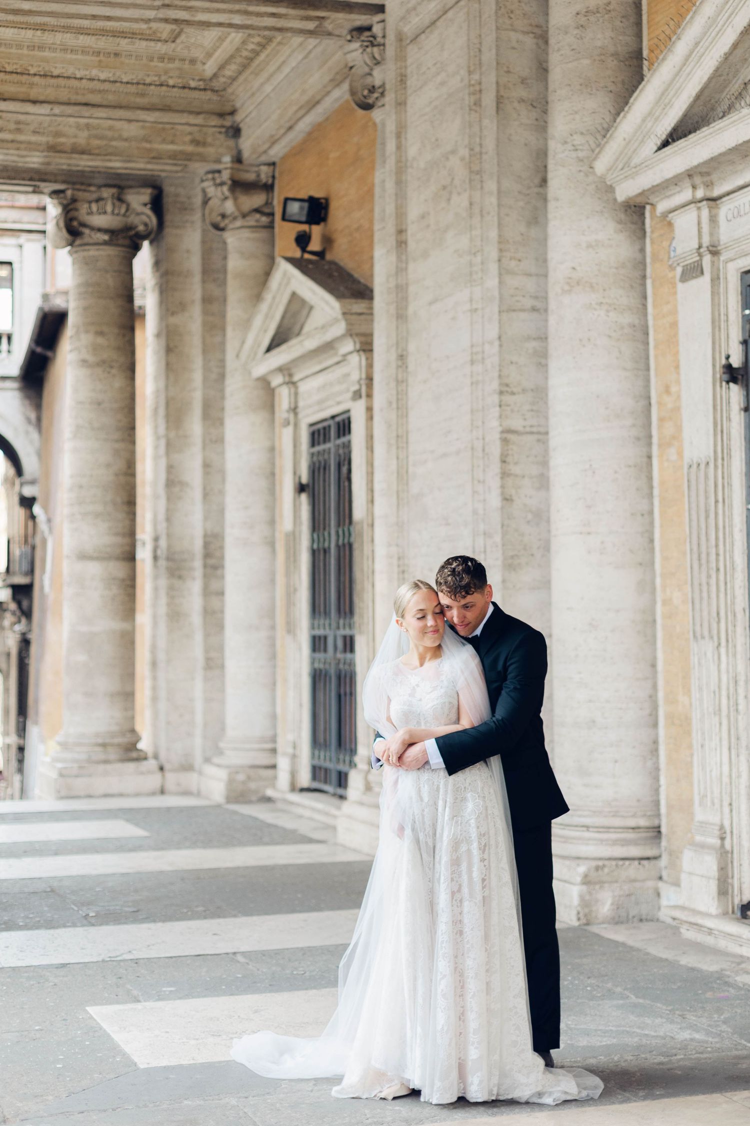 Newlyweds share an intimate moment under a grand stone colonnade with classical details of rome itlay.