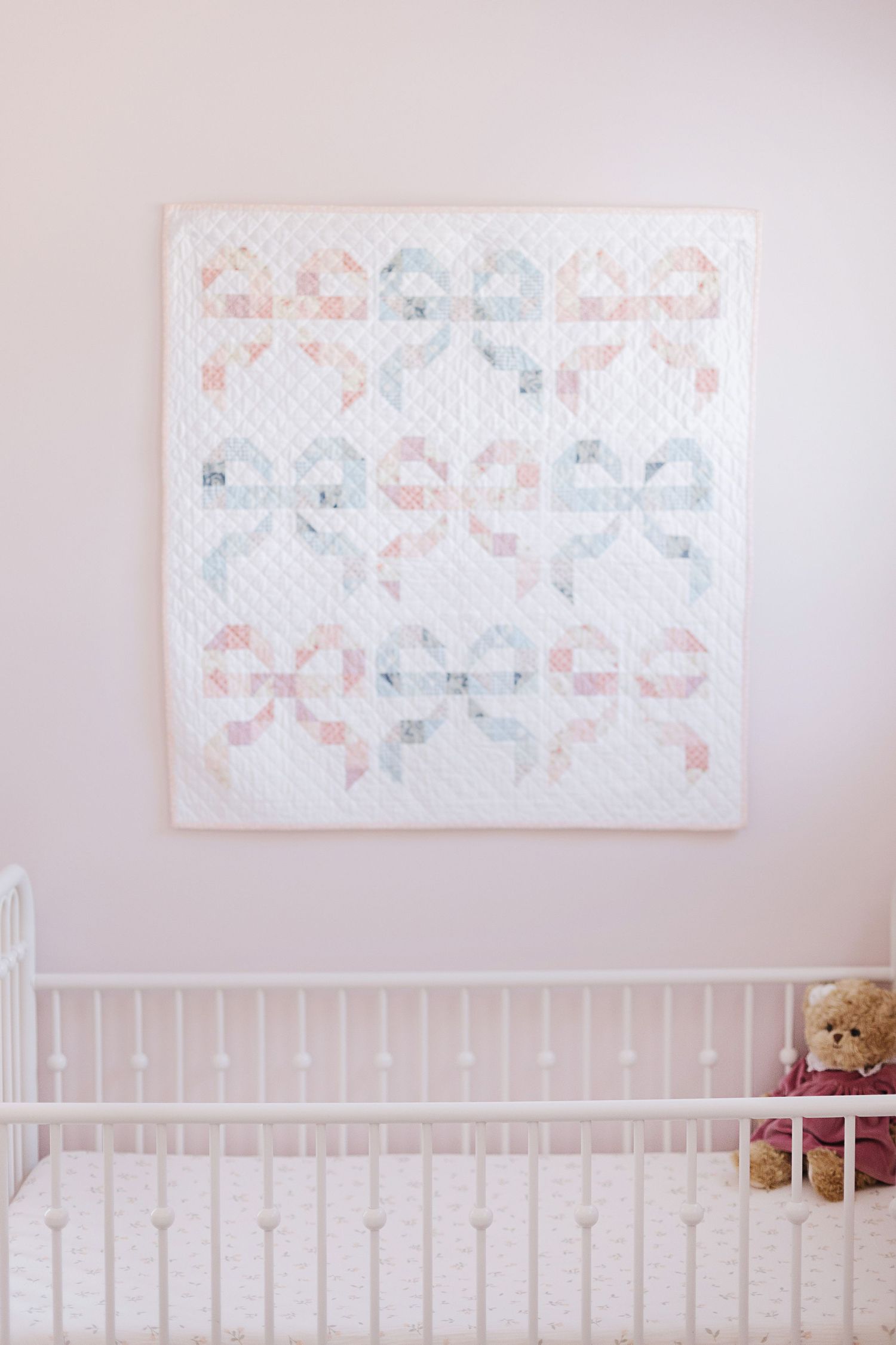 A pastel quilt pattern artwork hangs above a white crib with a teddy bear.