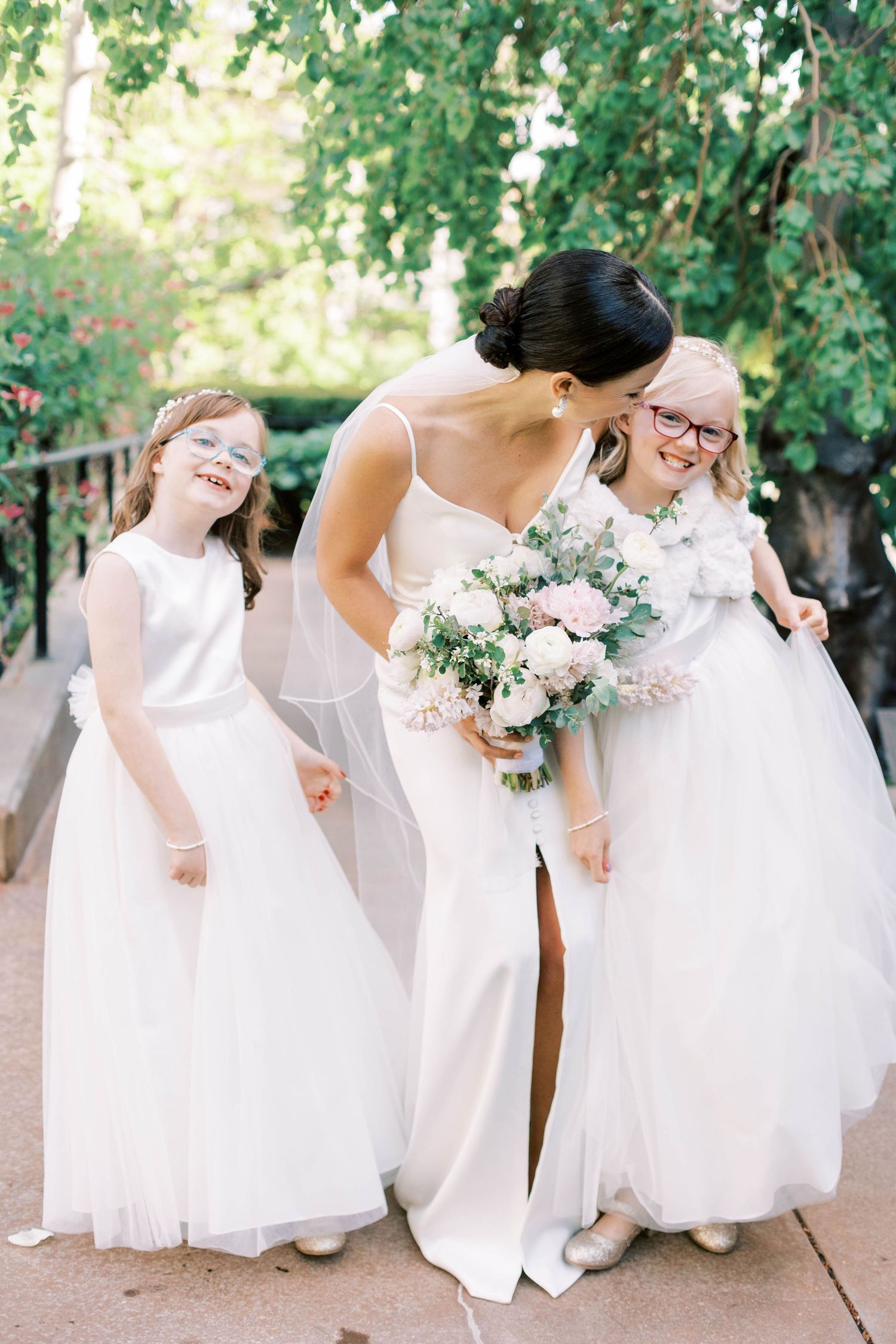 Three figures in flowing white gowns share a joyful moment while holding a pink and white floral bouquet.
