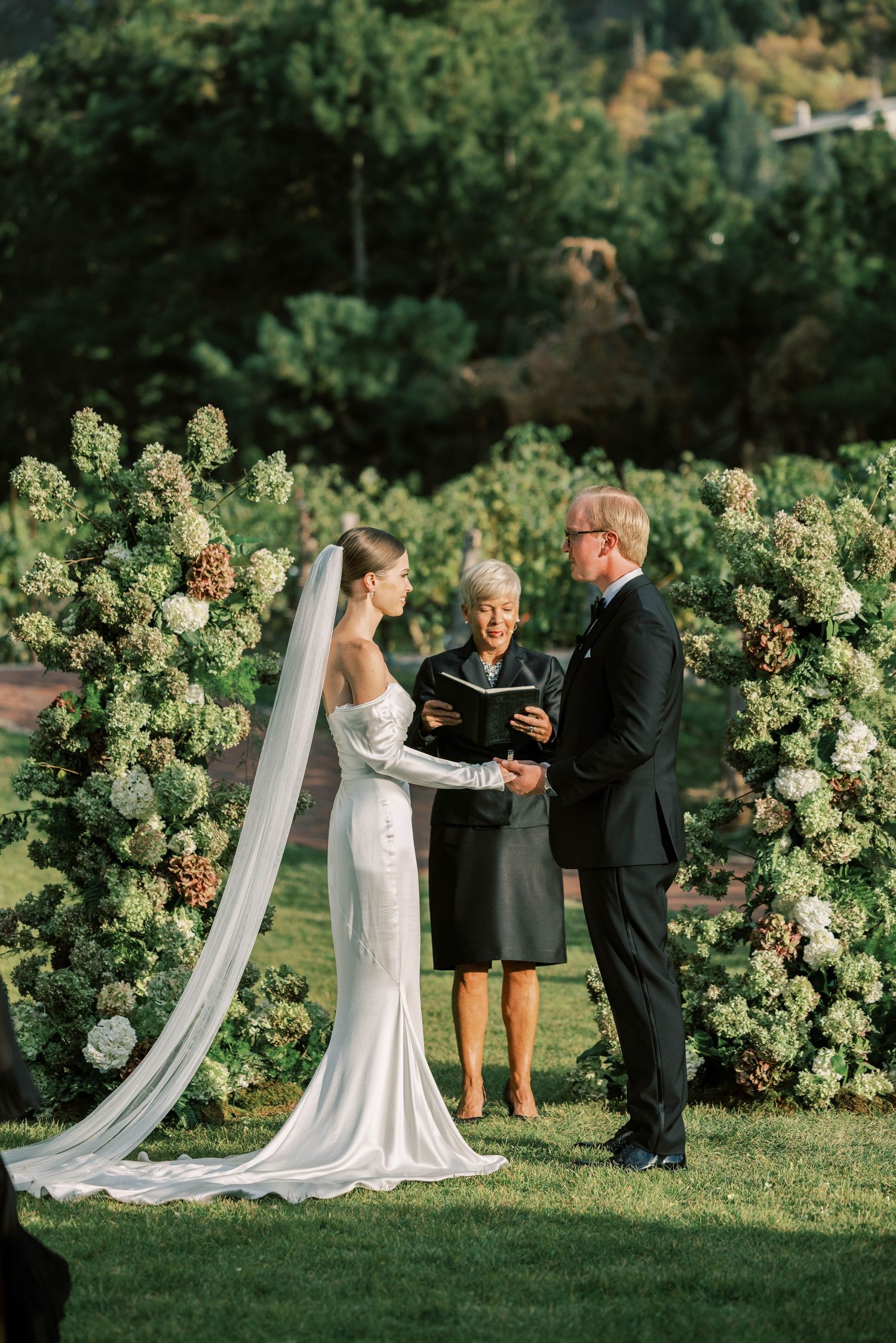 Outdoor wedding ceremony with floral arch and flowing veil in a garden setting with mountain views.