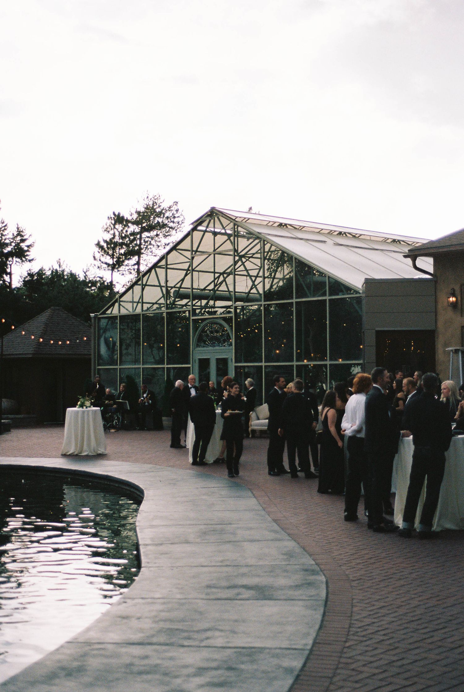 Modern glass greenhouse venue with guests mingling by a reflecting pool during an evening event.