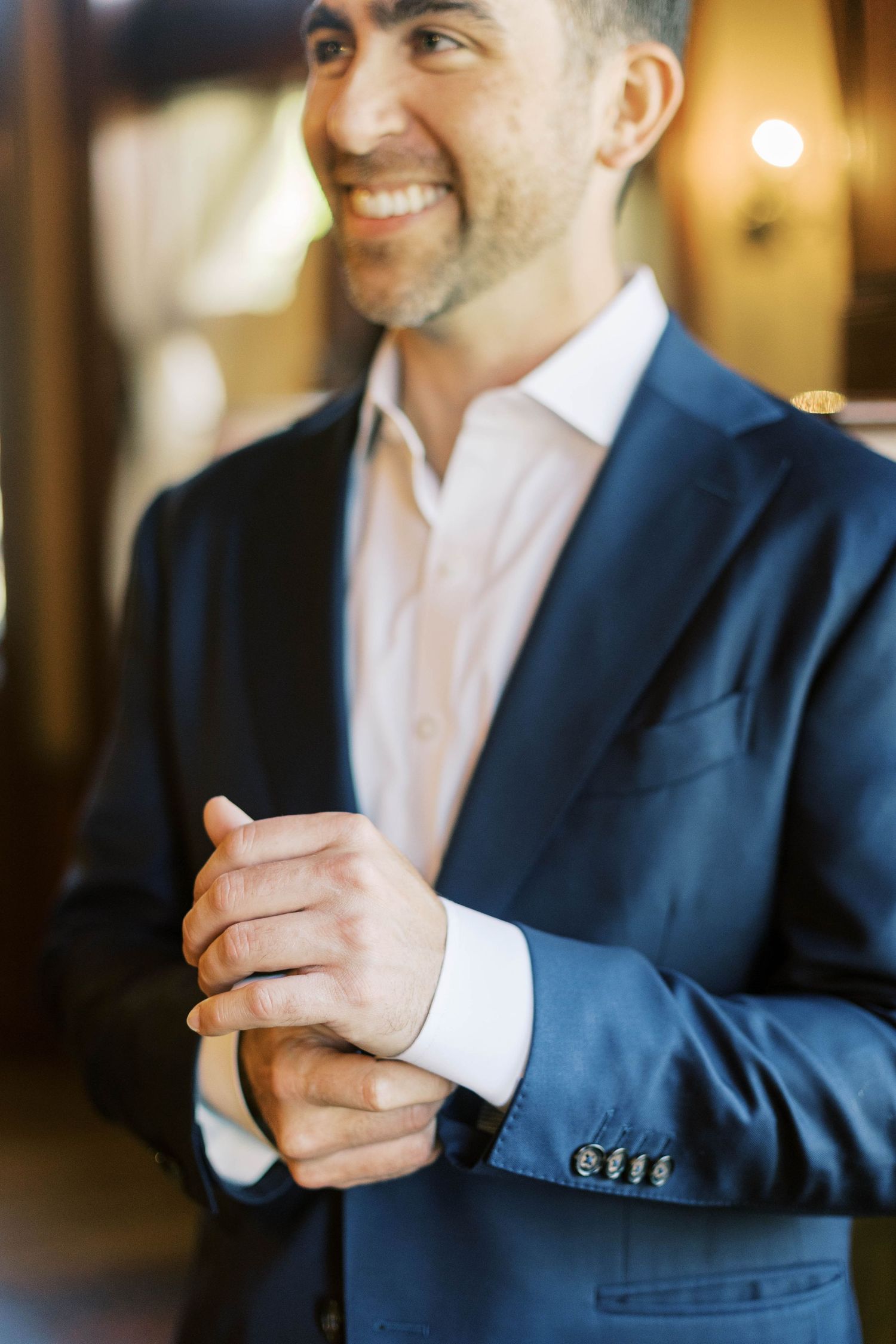 A person wearing a navy blue suit and pink dress shirt adjusts their cuffs with a warm smile.