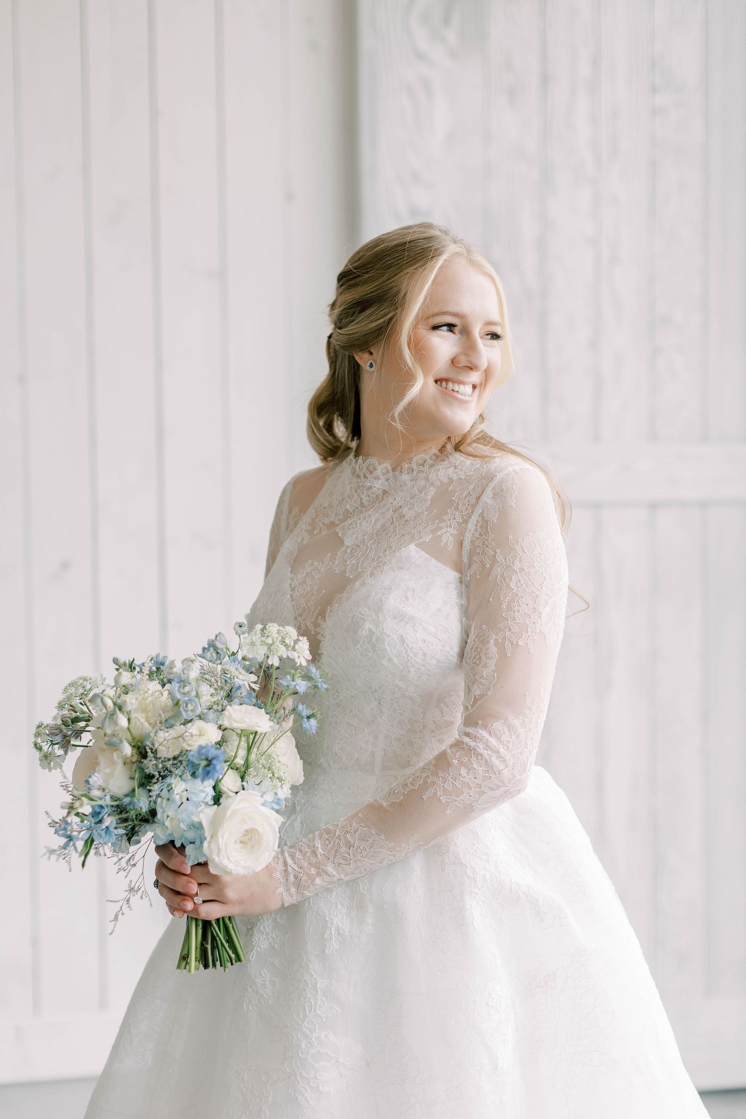Radiant bride in long-sleeve lace wedding gown holding blue and white floral bouquet.