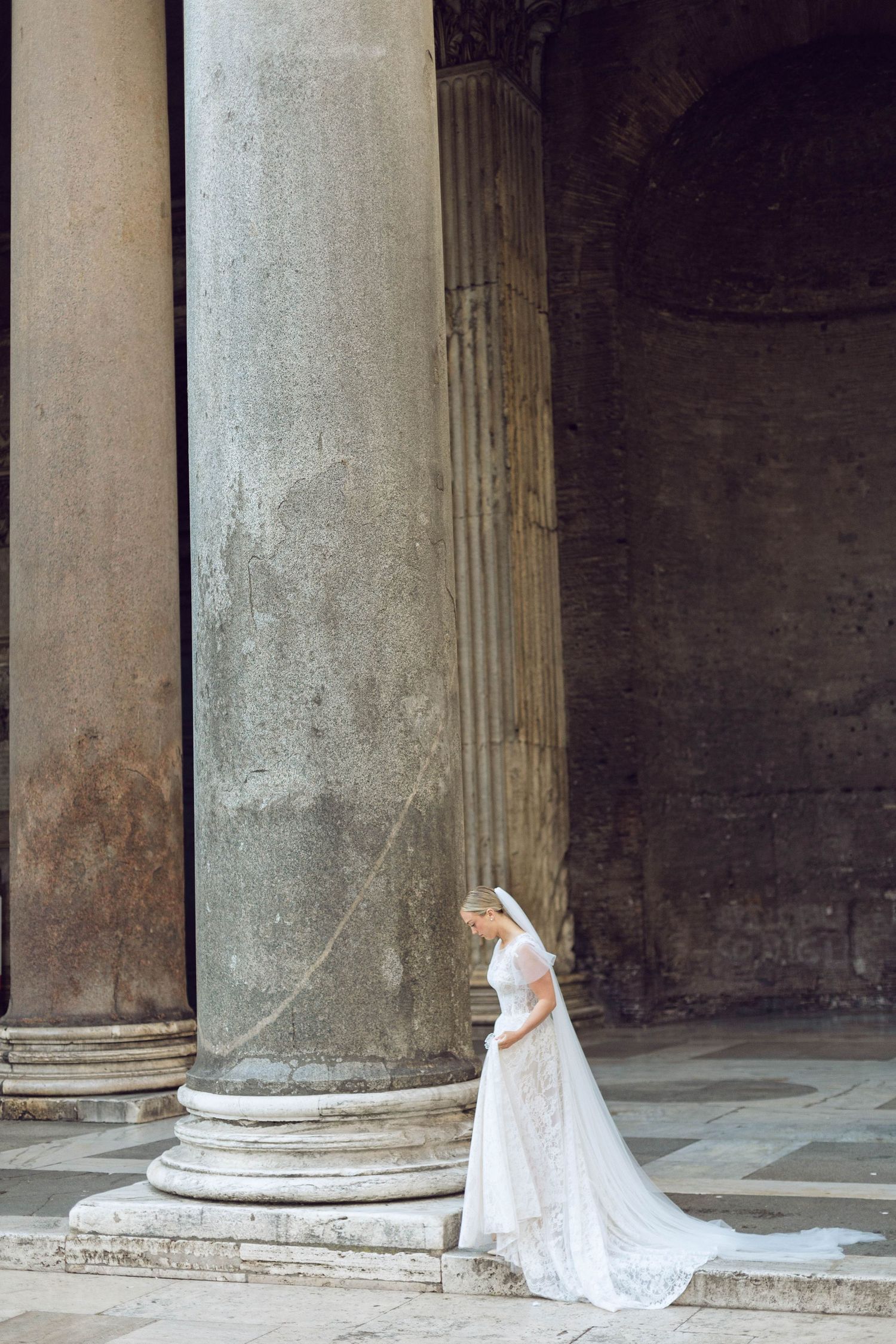 A wedding dress with long train flows elegantly between ancient stone pillars of the pantheon rome italy