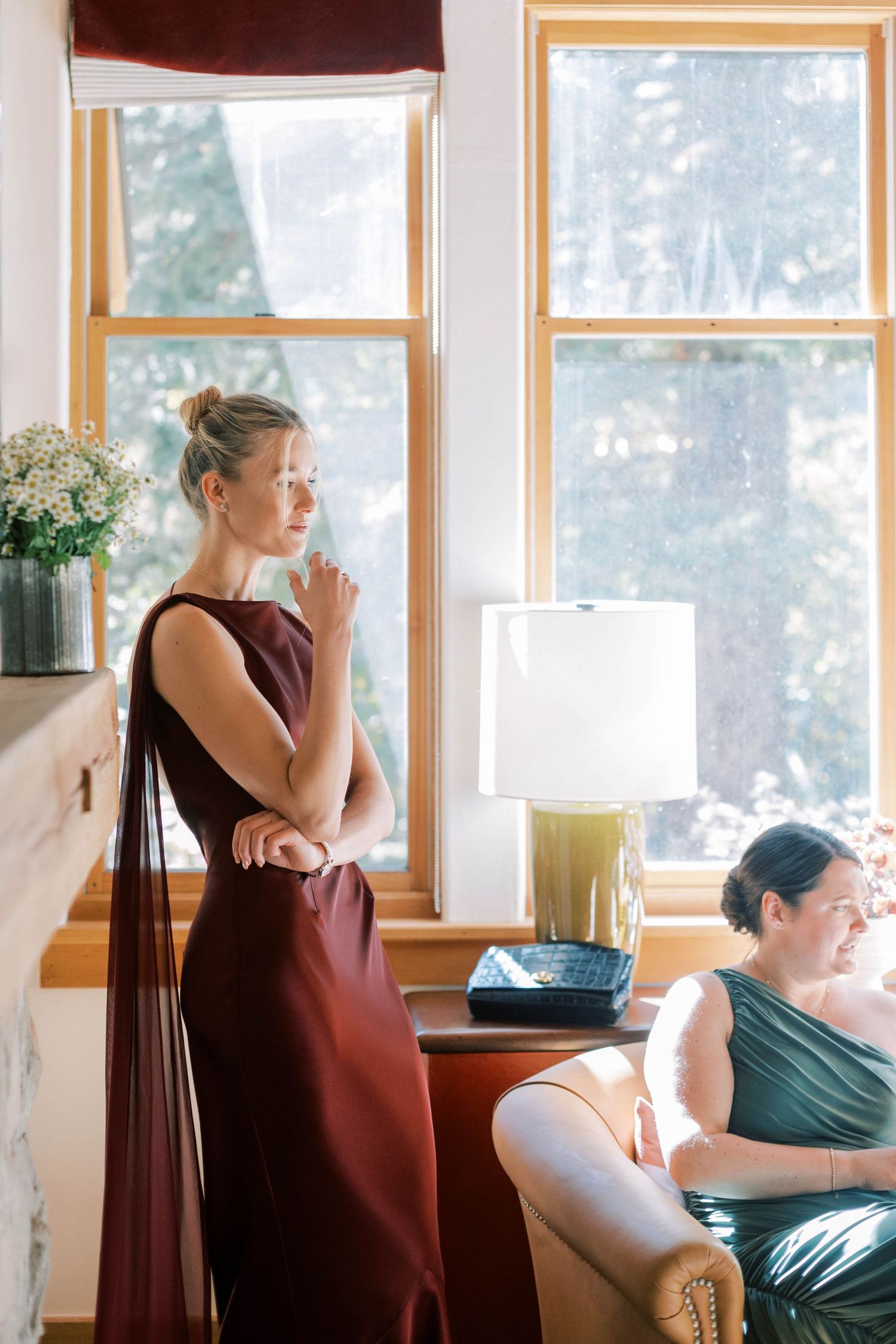 Two people in elegant dresses stand near a bright window with white curtains.