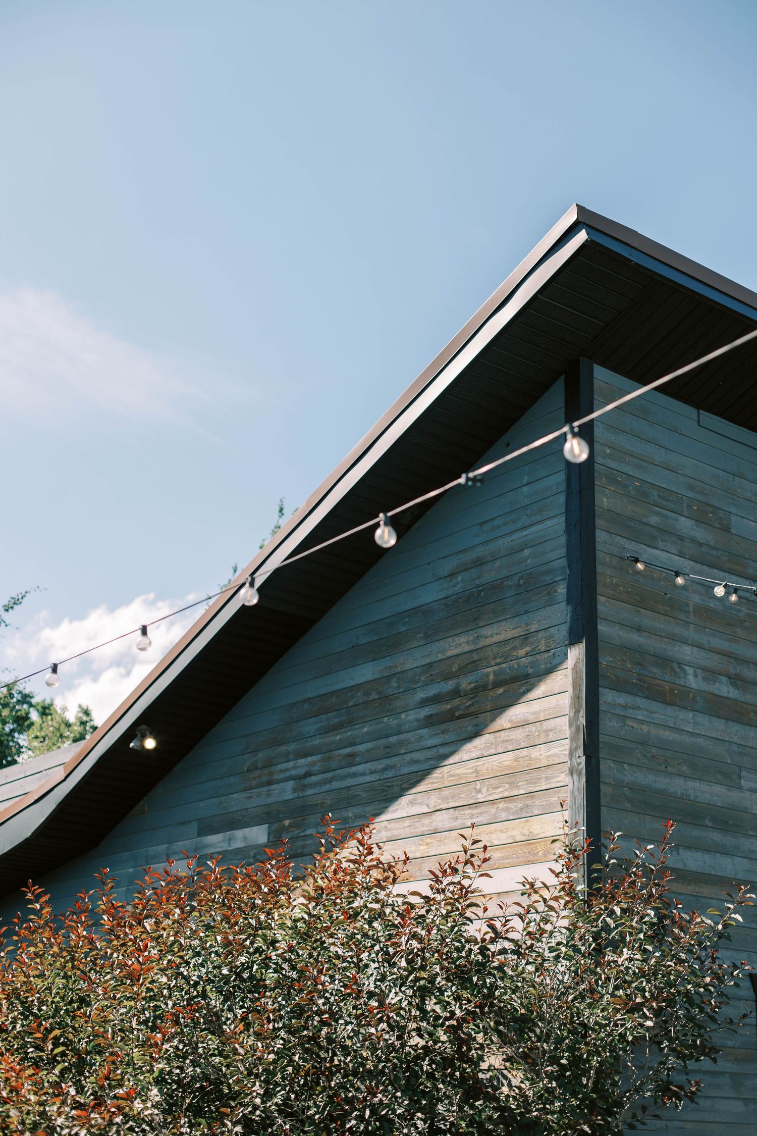 Modern rustic barn exterior with string lights and weathered wood siding against blue sky.