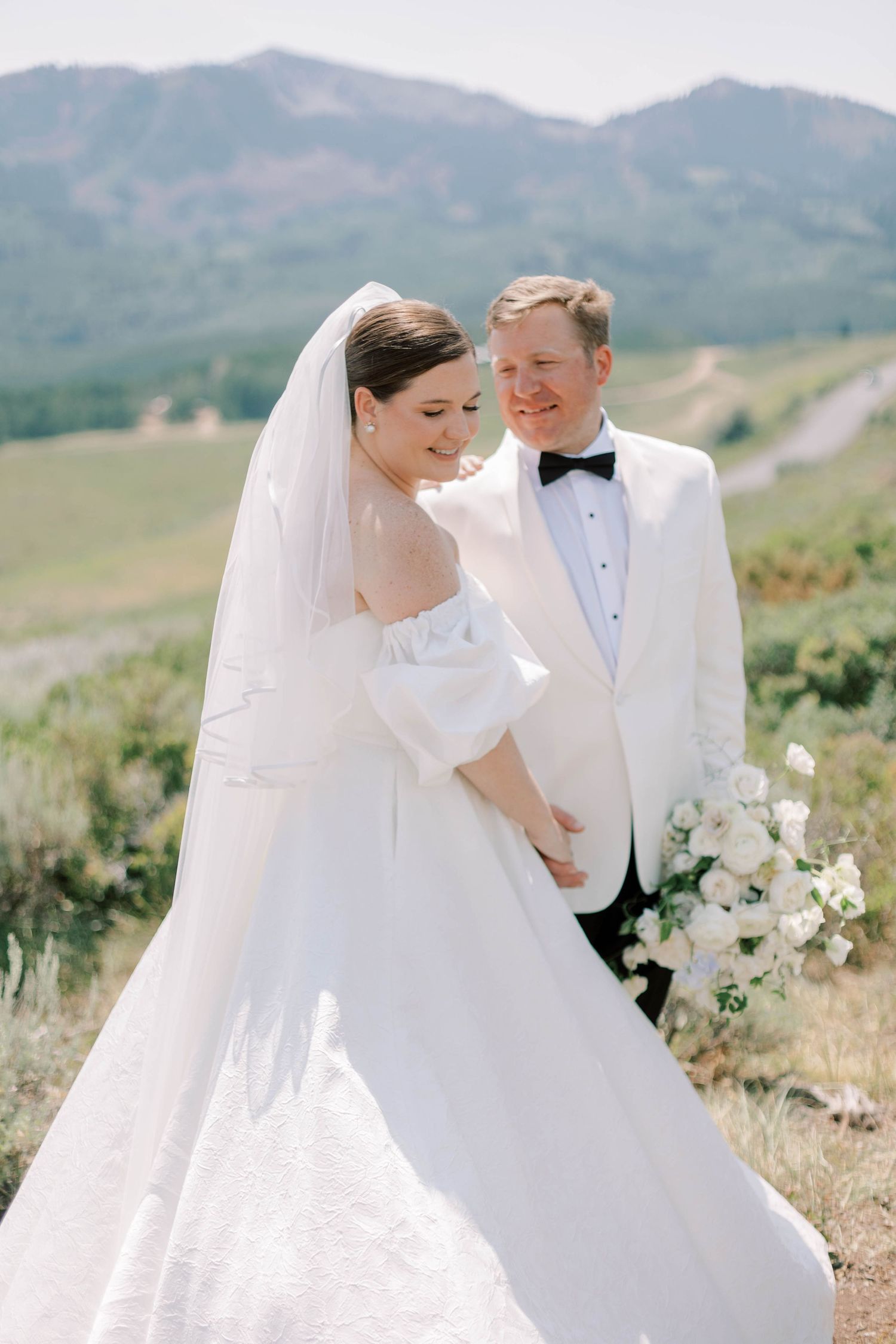 Wedding couple poses on a hillside with mountains, wearing white formal attire and holding a bouquet.