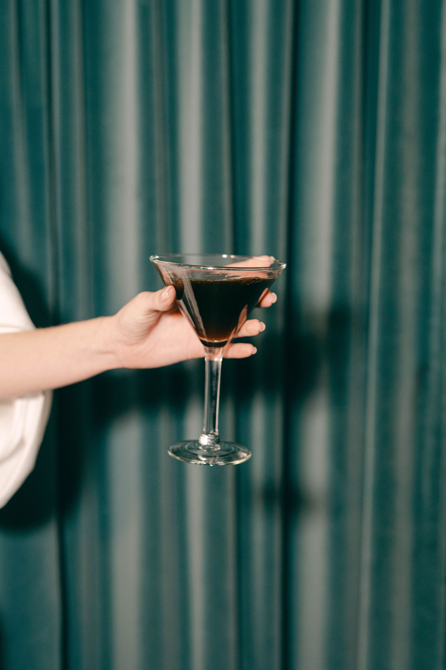 Hand holding a dark cocktail in martini glass against emerald green curtain backdrop.